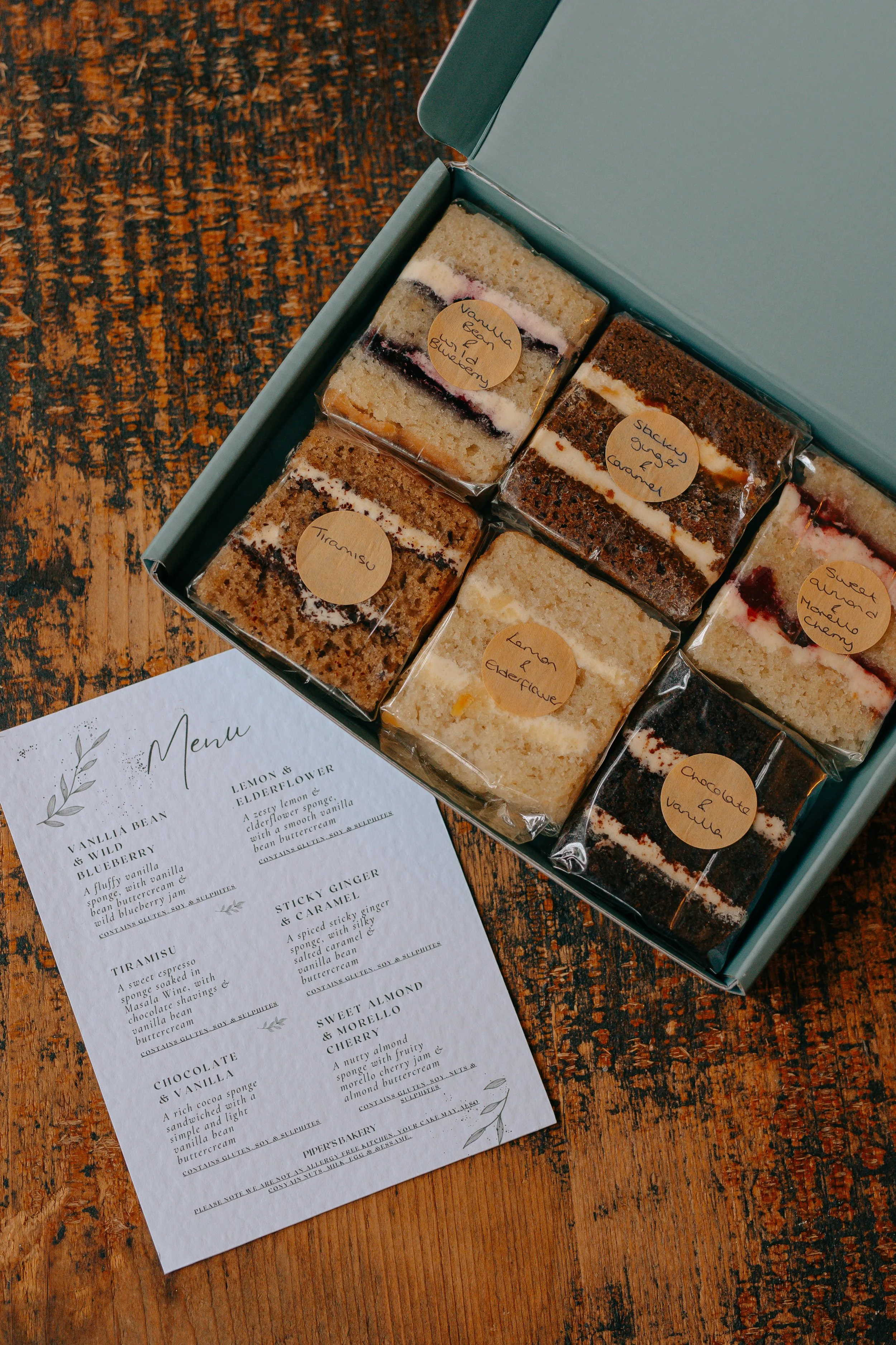 A sage green box containing six different vegan wedding cake flavours, including a menu, ready to be posted
