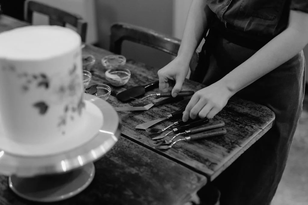 Cake designer arranging cake decorating tools on a rustic wooden table with a partially decorated cake in the foreground.