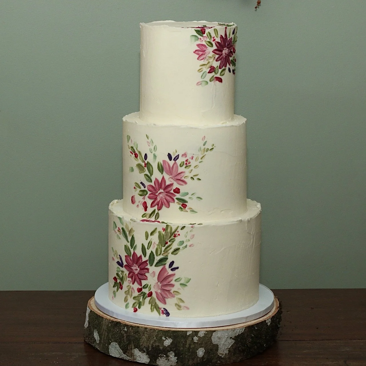 A three tier wedding cake with pink and green hand-painted flowers on the left side and the top right and rustic white icing
