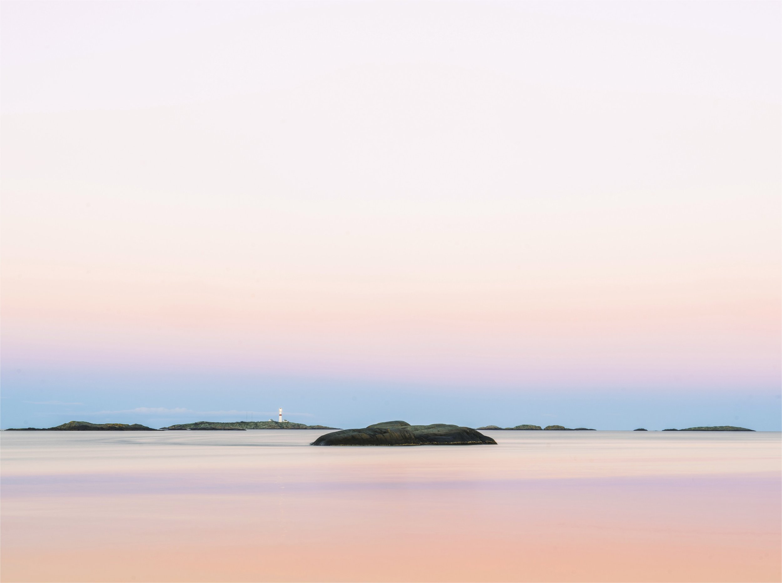 Calm sea with distant islands, rocks in the foreground, lighthouse on one island, pastel-colored sky