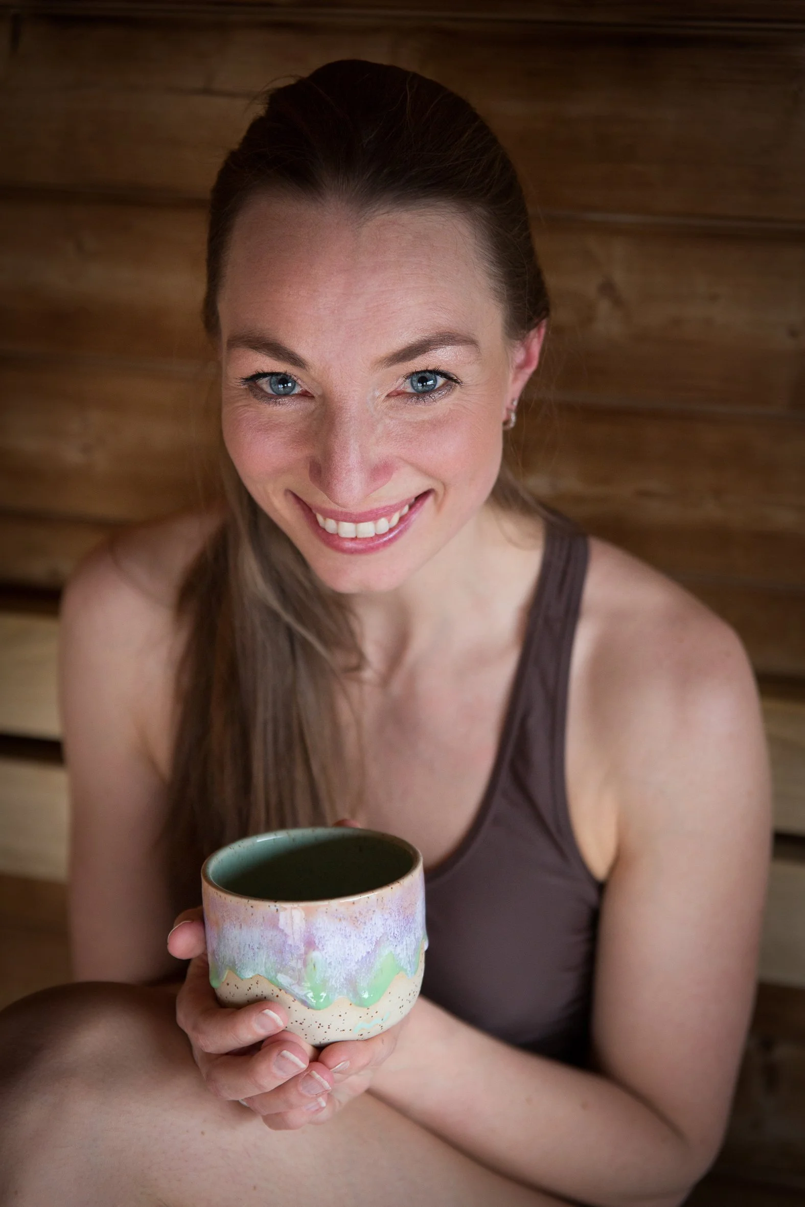 A smiling woman with blue eyes and long brown hair, wearing a brown sleeveless top, holding a colorful ceramic mug with a dark liquid, sitting in front of a wooden wall.