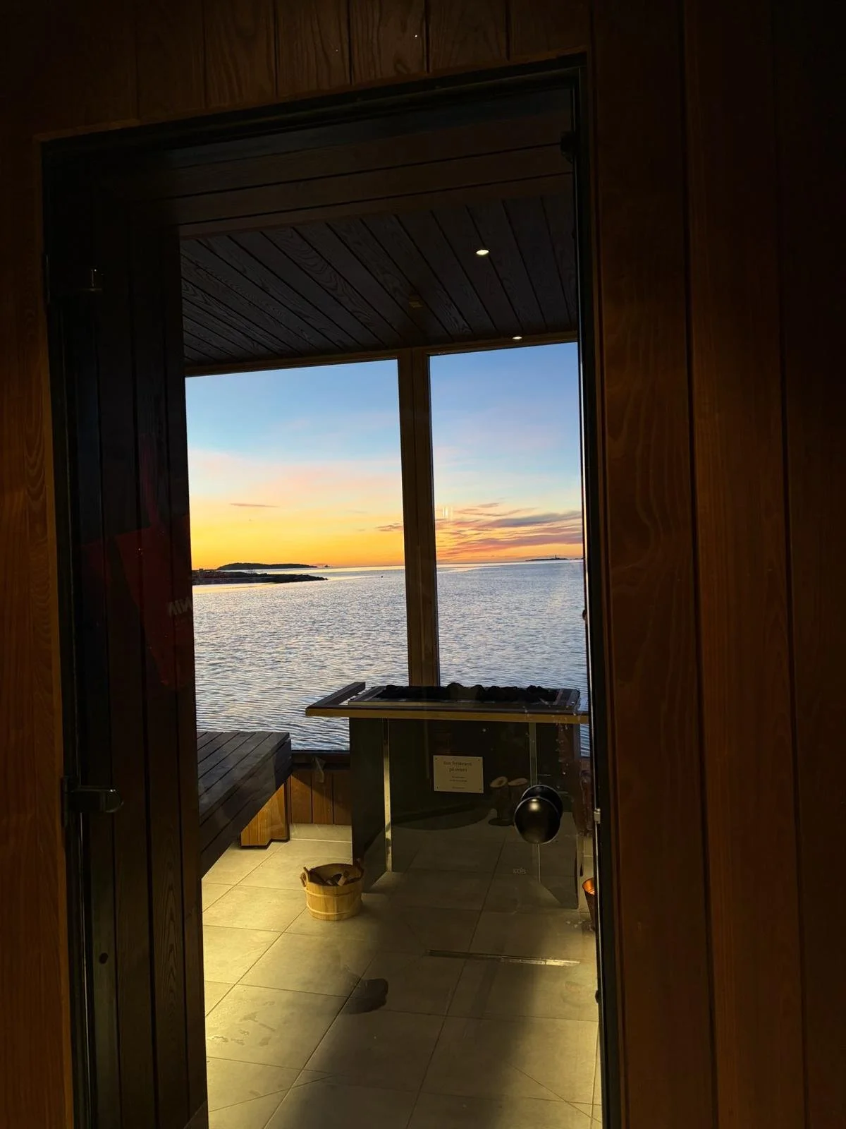 Et rom med store vinduer som viser en solnedgang over havet, innredet med et badestamp, benker og en liten høyde ved vinduet.