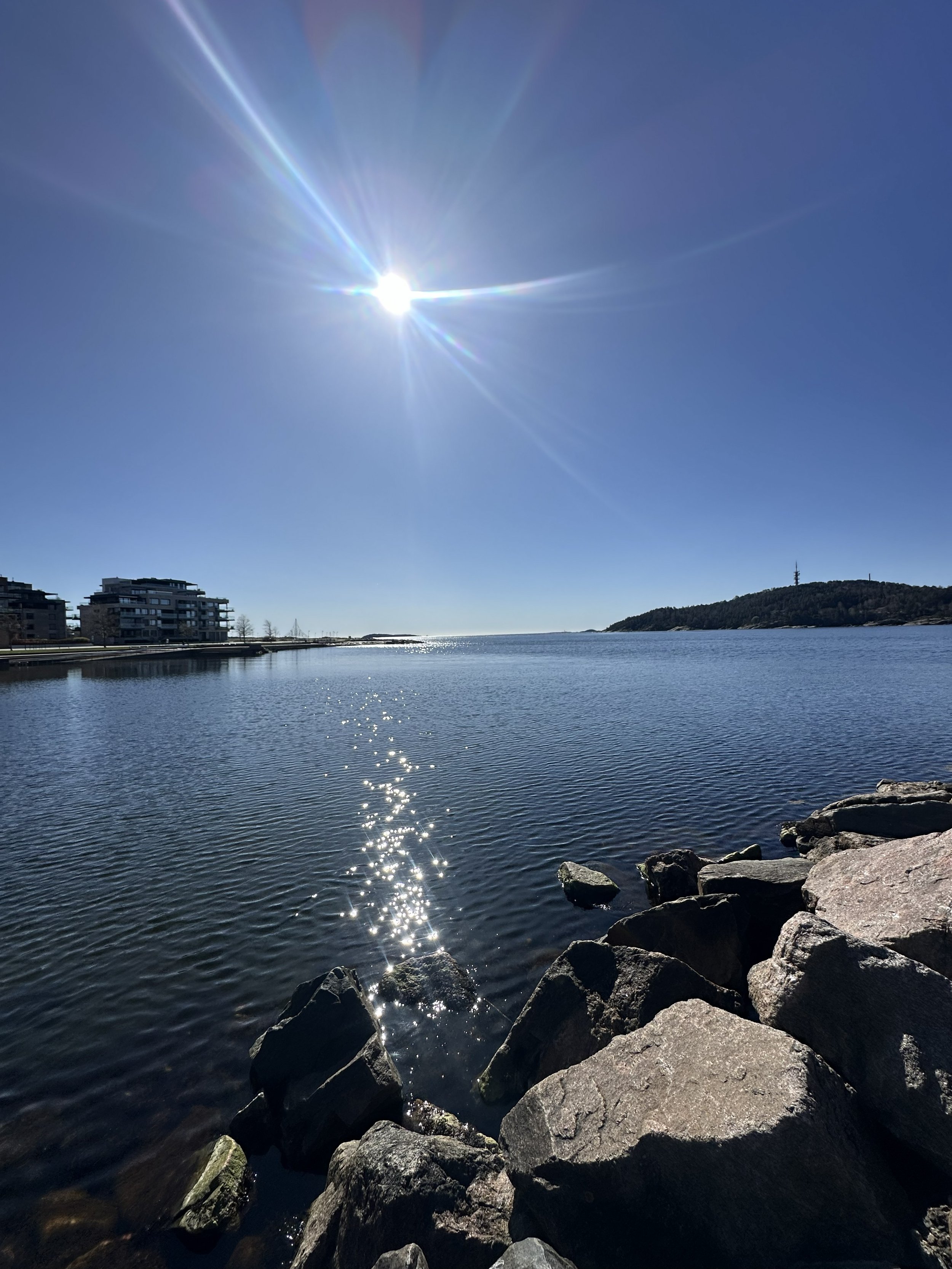 Sun shining brightly over a calm body of water with rocks along the shoreline and buildings visible in the background. A hill is seen in the distance.