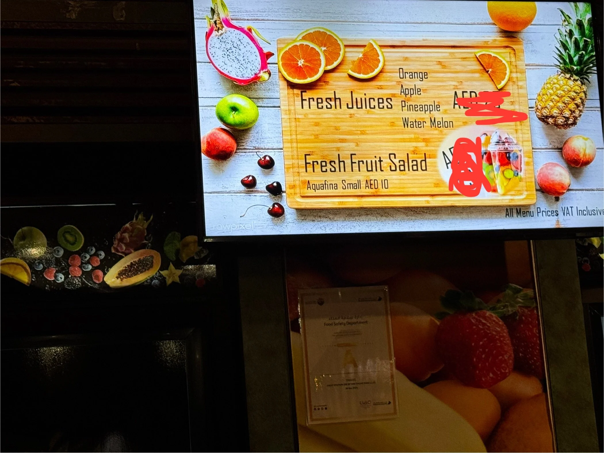 Menu board displaying fresh juices and fruit salad options with images of fruits like dragon fruit, oranges, apples, and a pineapple. The board includes prices in AED, and the mention of Aquafina bottled water availability.