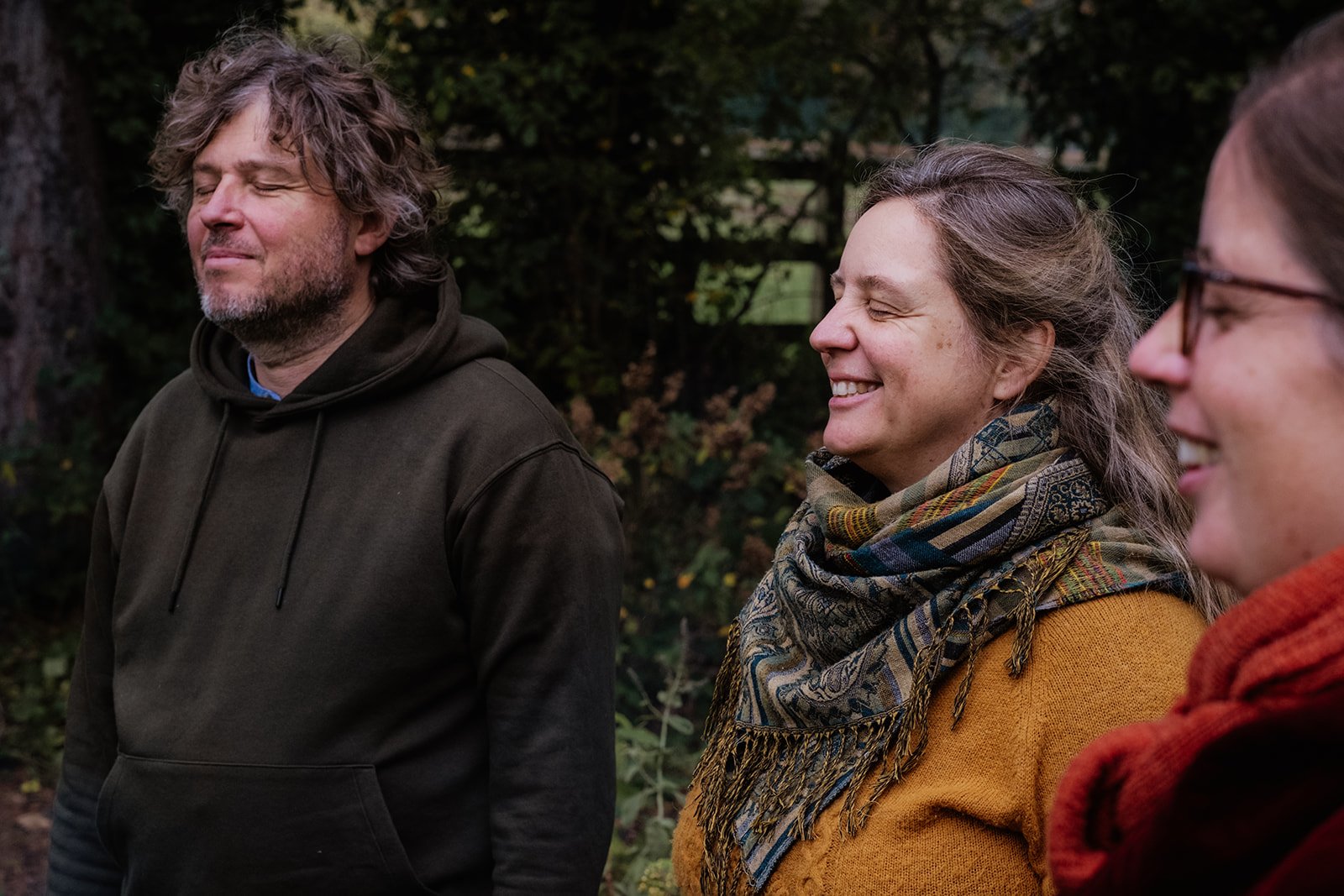 Drie mensen staan buiten, lachen en genieten met gesloten ogen van het moment. De achtergrond is een groene, bosachtige omgeving.