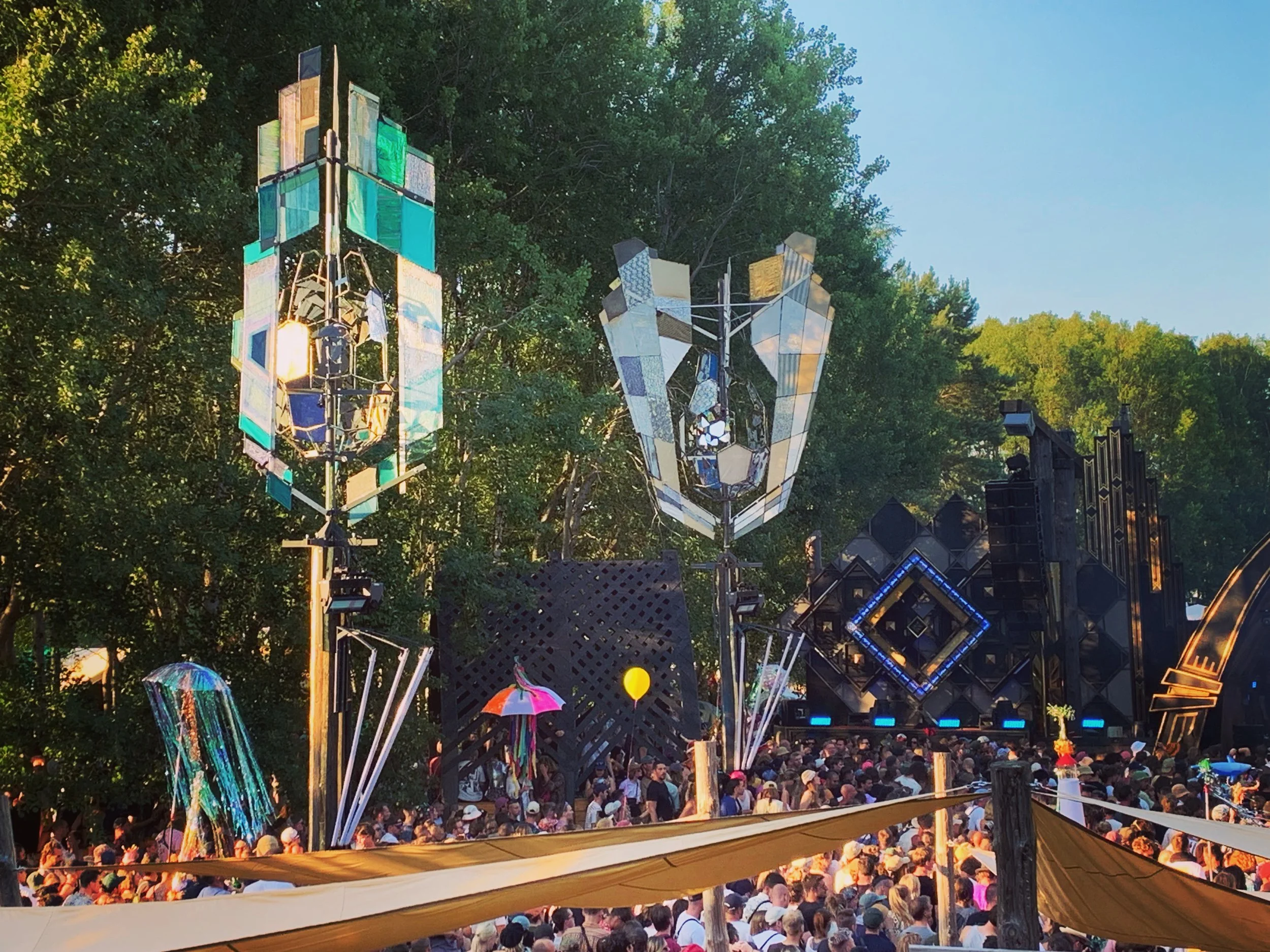 Festivalszene im Freien, mit bunten Leuchttürmen, einem großen Publikum und einer Bühne im Hintergrund, umgeben von Bäumen bei Sonnenuntergang.