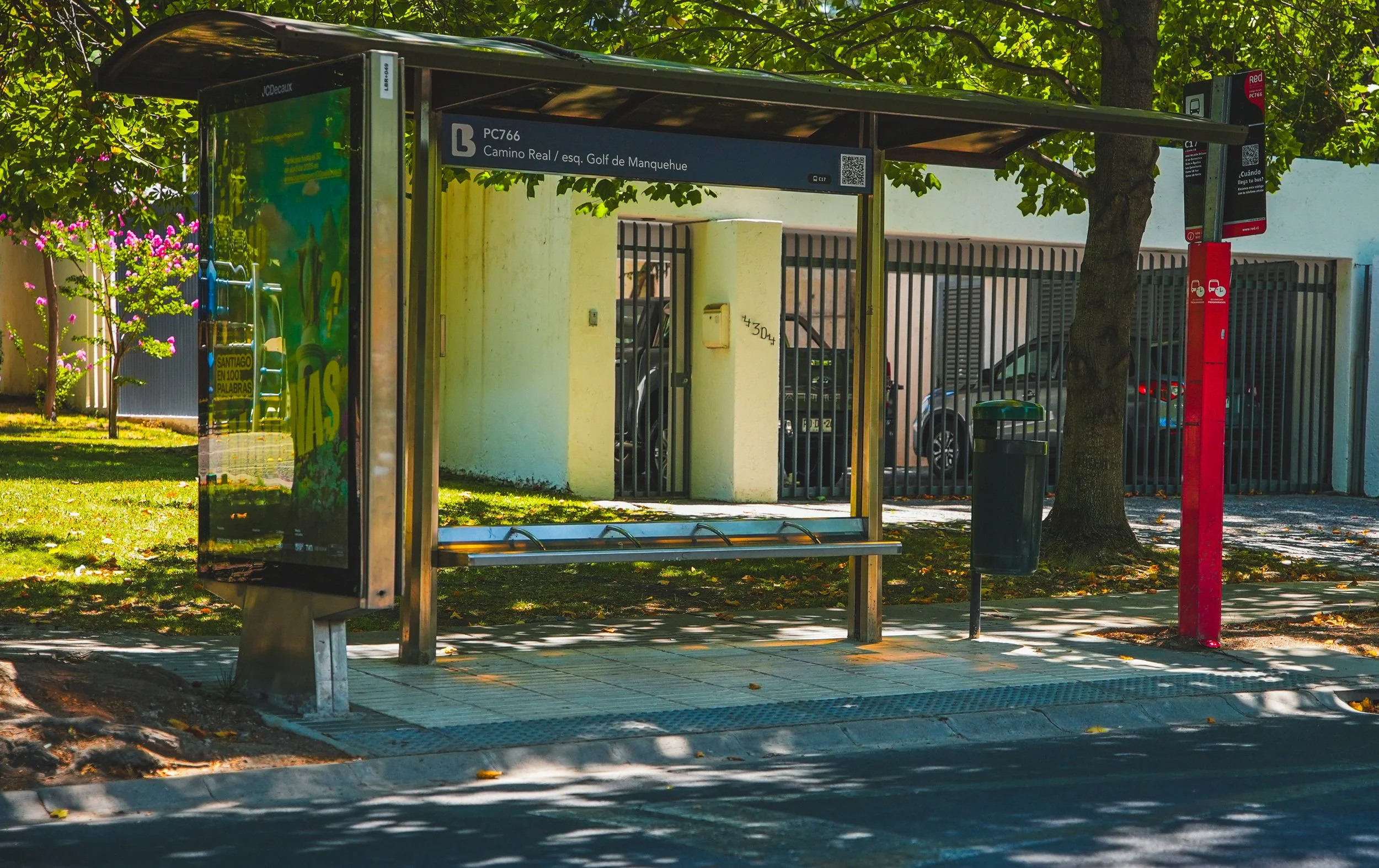 Parada de autobús en un lugar sombreado con advertencias para subir al autobús y un teléfono móvil. Hay arboles y coches en el fondo.