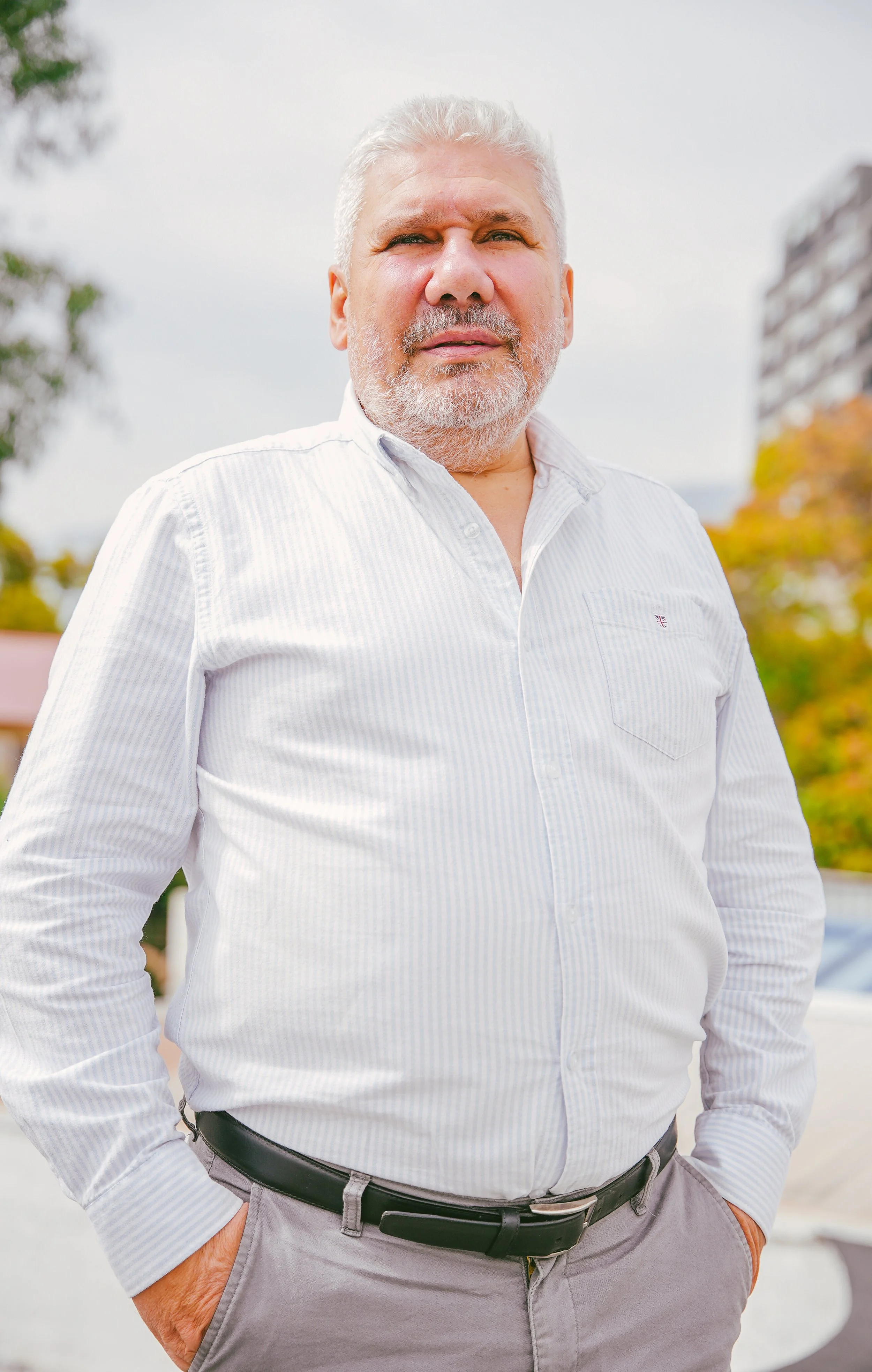Hombre con cabello gris y barba, viste camisa blanca a rayas y pantalones grises, posa con las manos en los bolsillos en un entorno urbano con árboles y edificios.