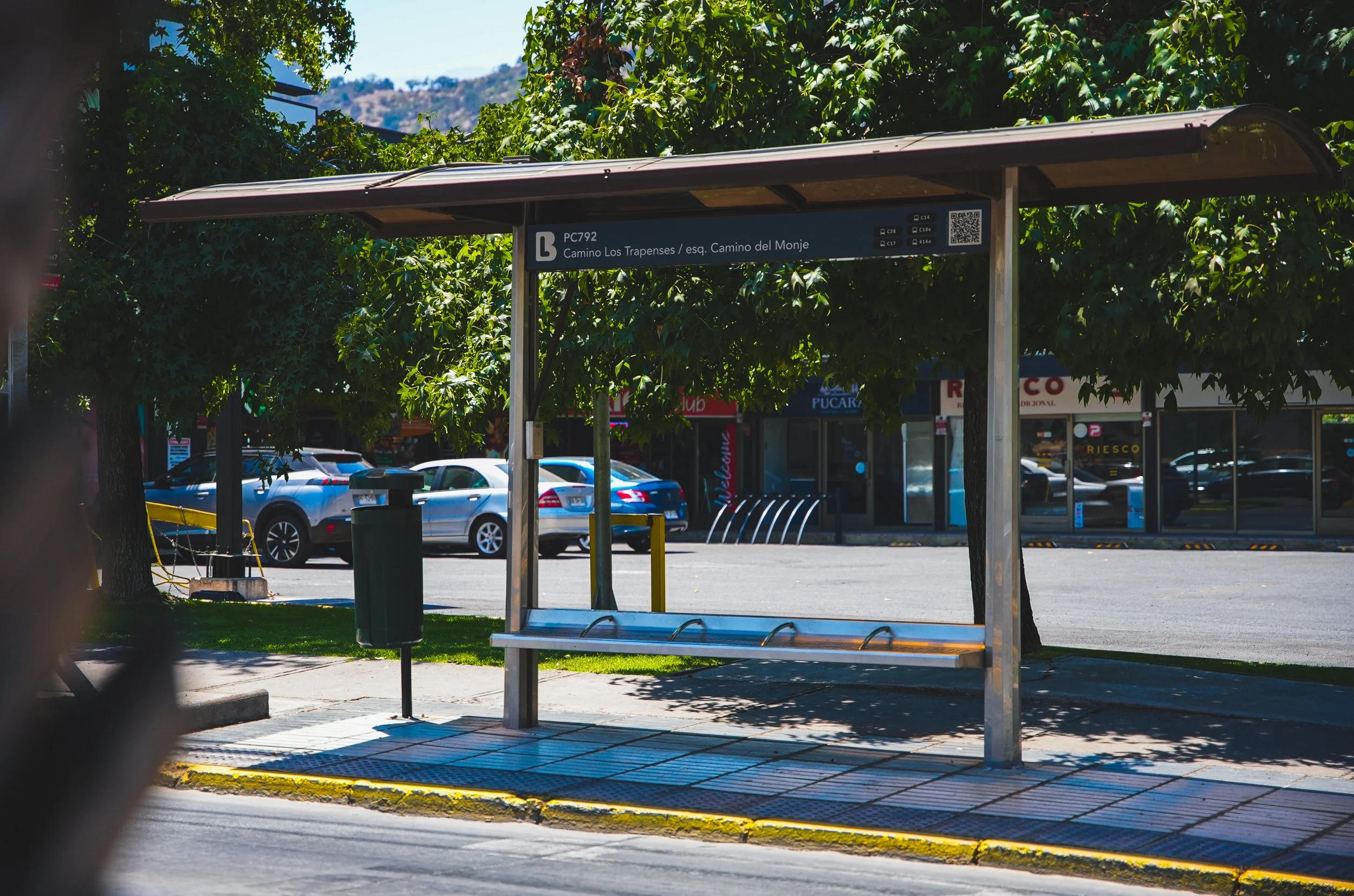 Parada de autobús con un techo y un cartel que indica la ruta PC792 en una calle con árboles y autos aparcados al fondo.