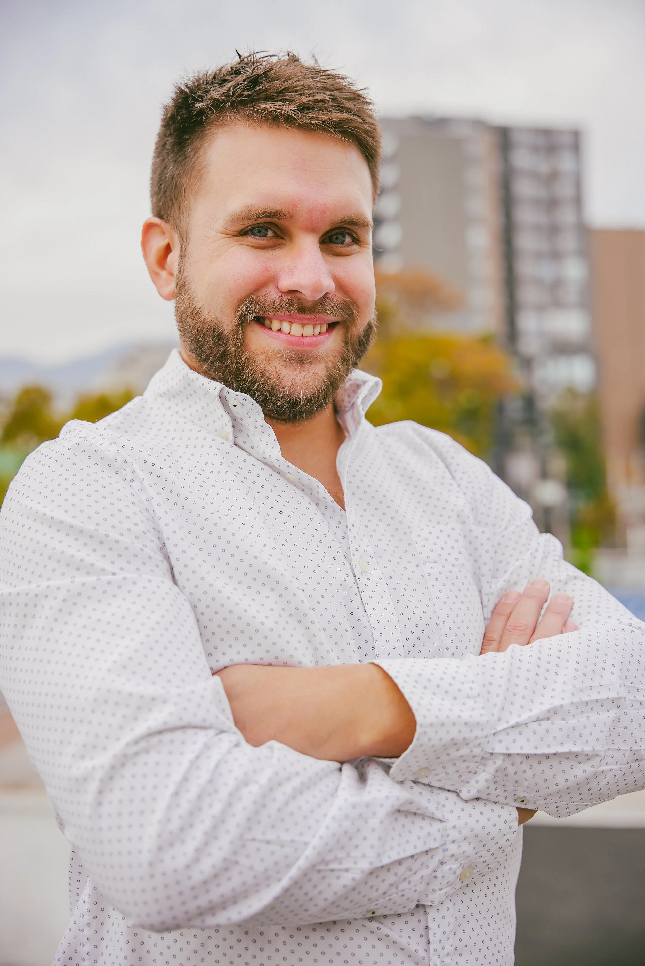 Hombre sonriendo con barba, piel clara, ojos verdes, cabello castaño, vistiendo camisa blanca con pequeños puntos, con los brazos cruzados, en un entorno urbano con edificios y árboles en el fondo.