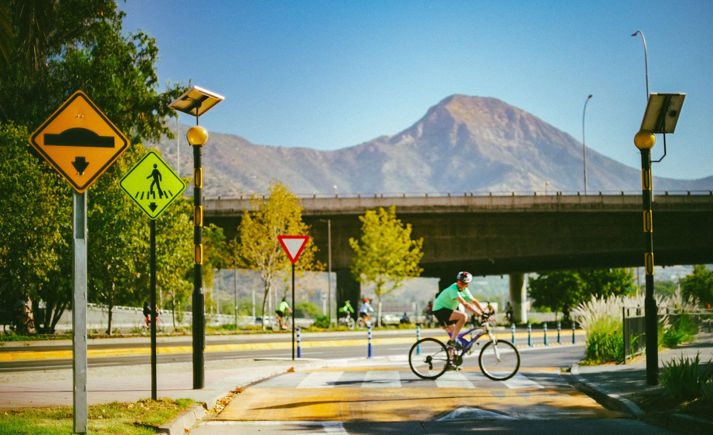 Una calle soleada con ciclistas y señales de tránsito, árboles y montañas al fondo.