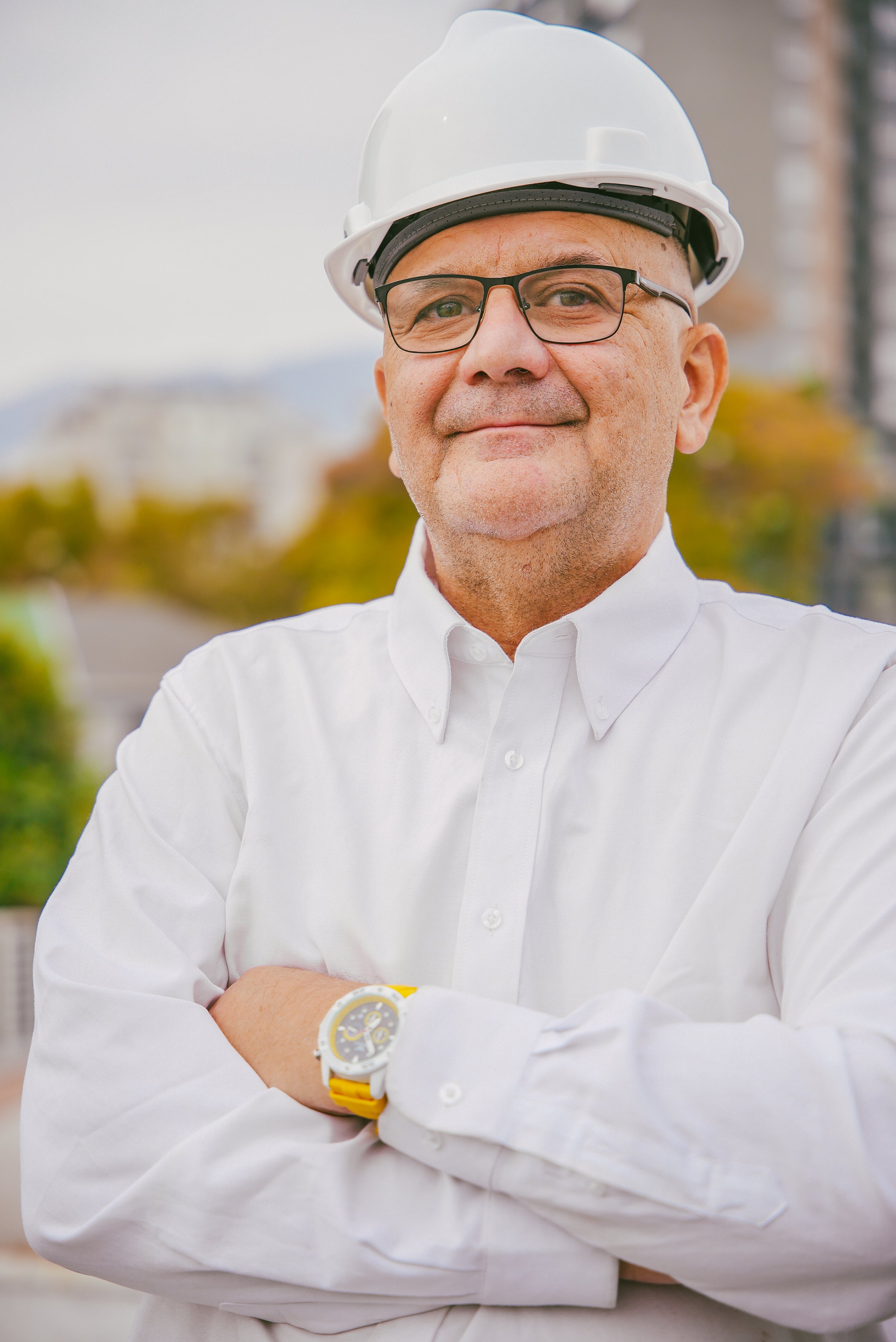 Hombre de mediana edad con gafas, usando casco de seguridad blanco y camisa blanca, con brazos cruzados y reloj amarillo, en un entorno urbano con edificios y árboles en el fondo.