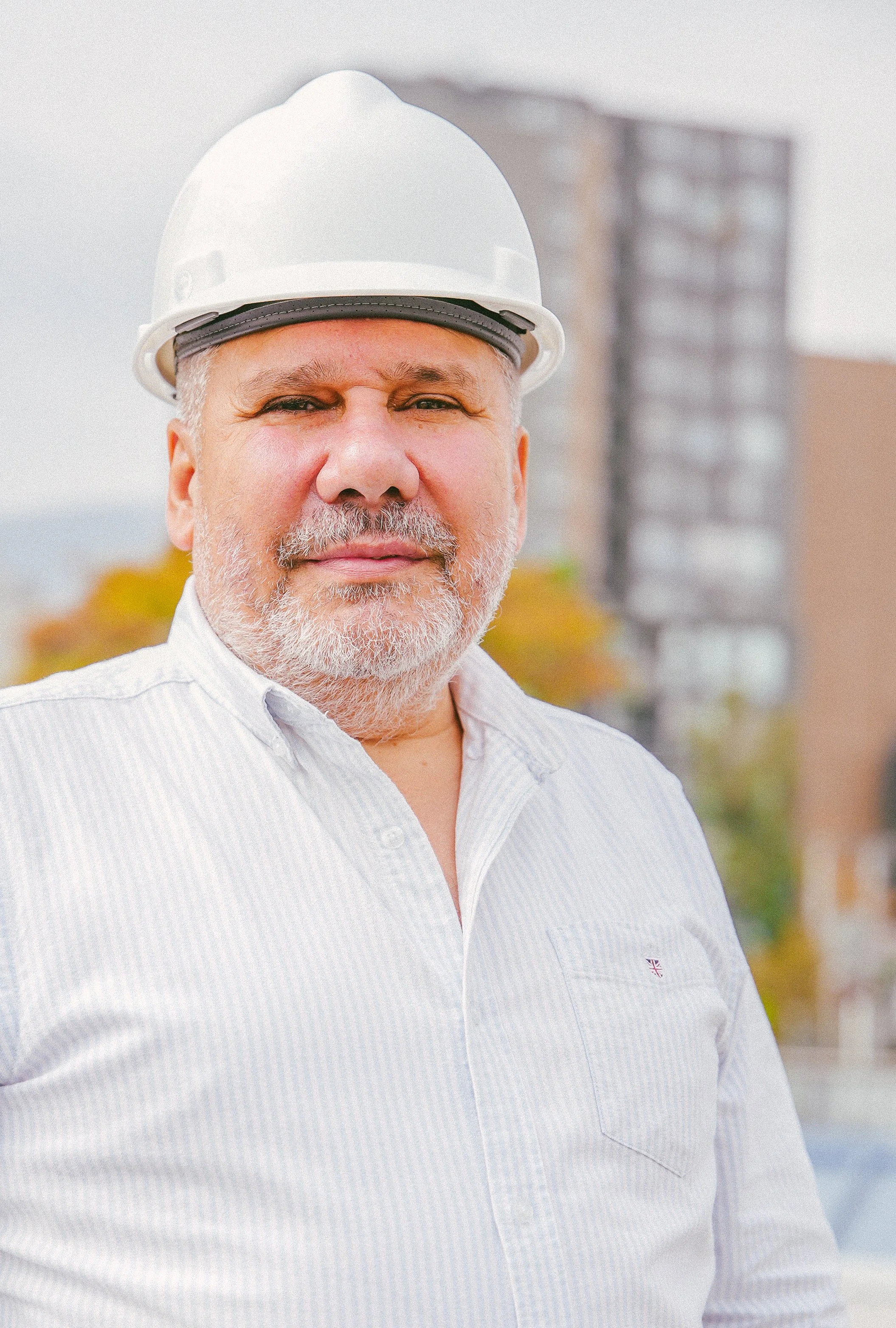 Hombre de mediana edad con barba canosa, usando casco de seguridad y camisa blanca, posando en un sitio de construcción con edificios en el fondo.