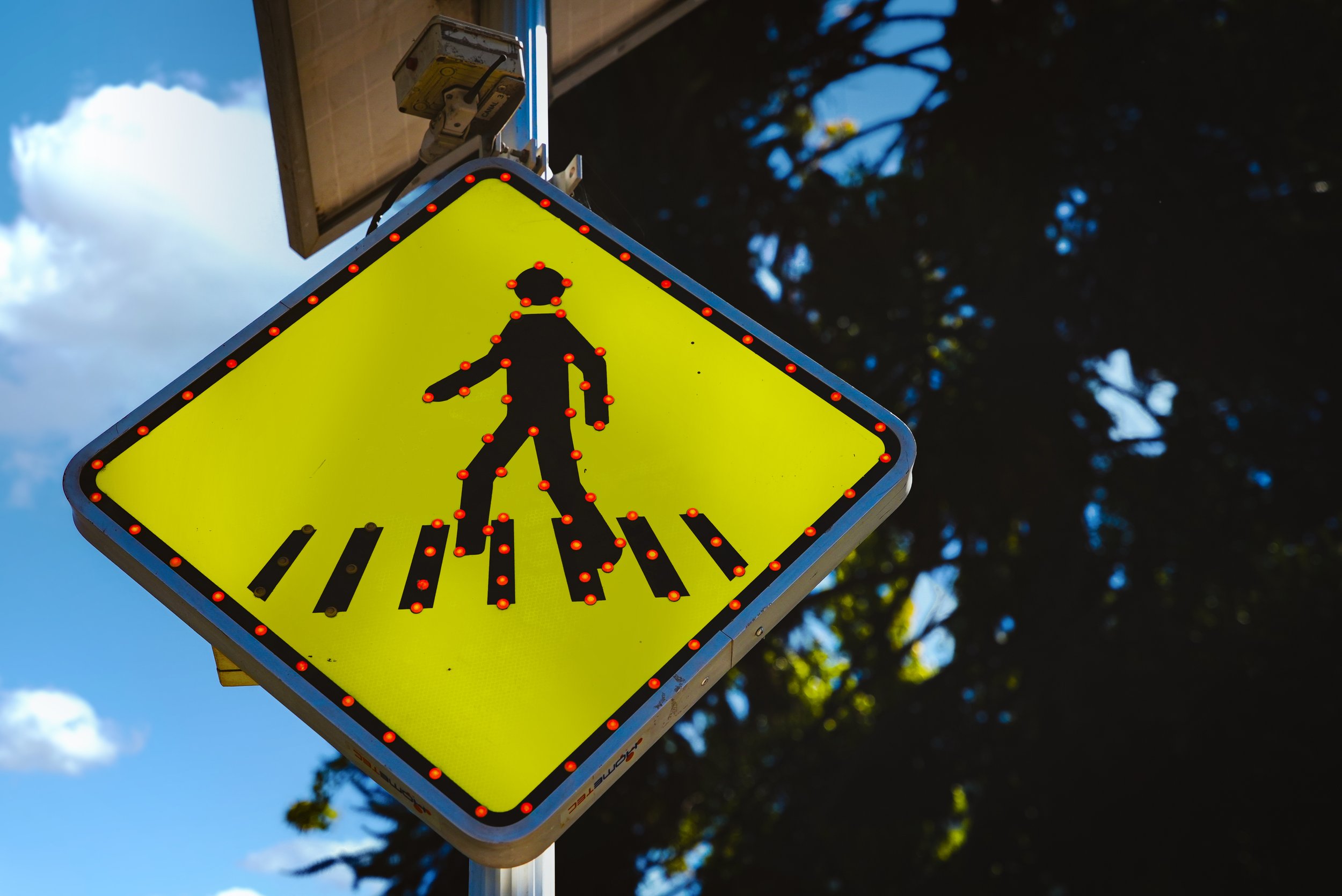 Señal de cruce peatonal iluminada con luces LED, con fondo de cielo y árboles.