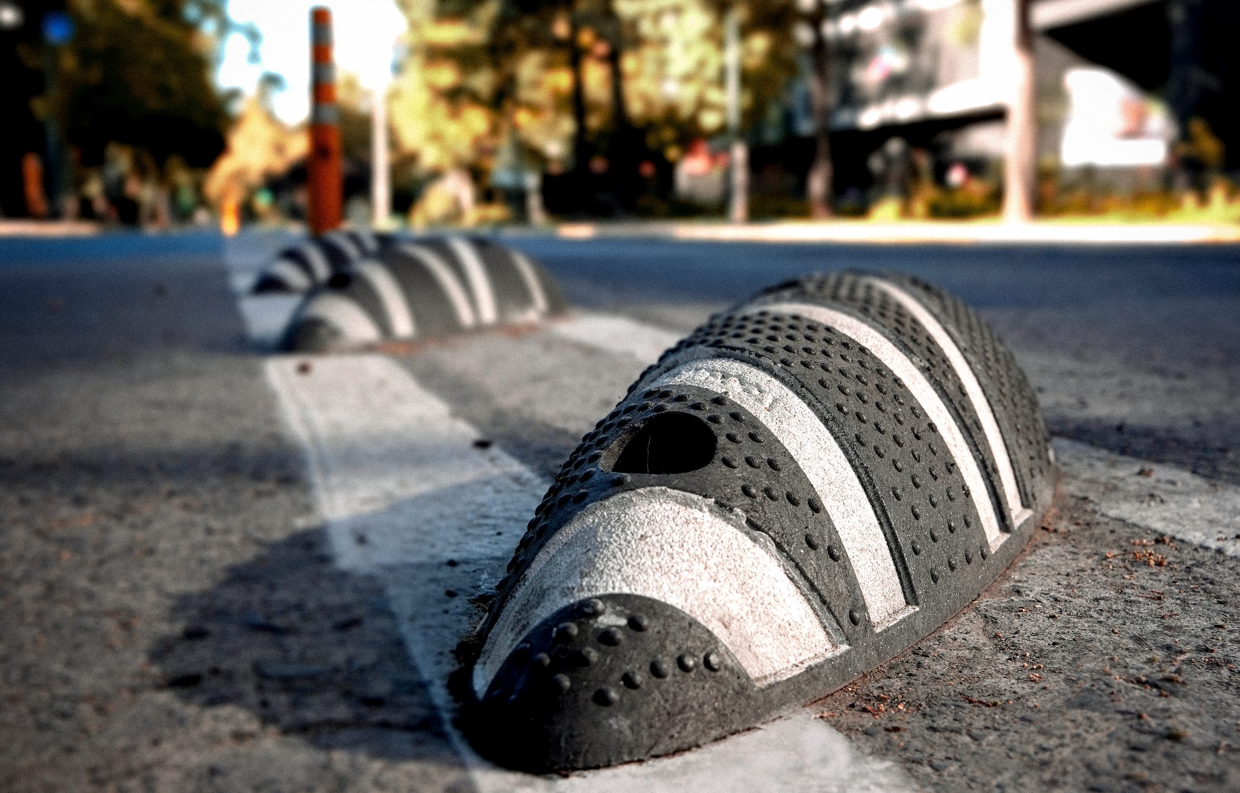 Barrera de protección en un paso peatonal en la calle, vista en primer plano, con un fondo difuso de árboles, edificios y una persona caminando.