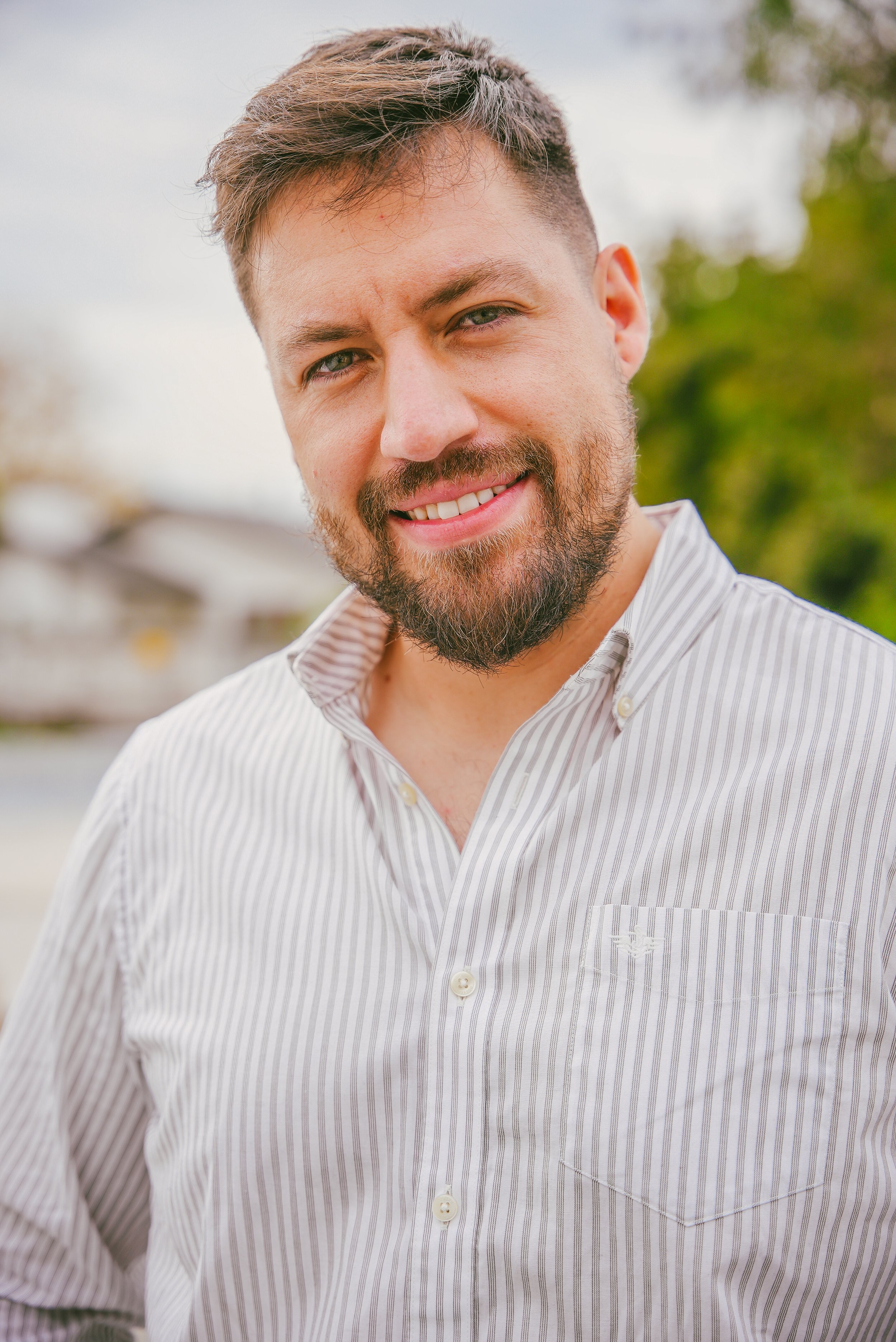 Retrato de un hombre con barba y cabello castaño, sonriendo, con una camisa de rayas blancas y grises, al aire libre con fondo de árboles y cielo nublado.