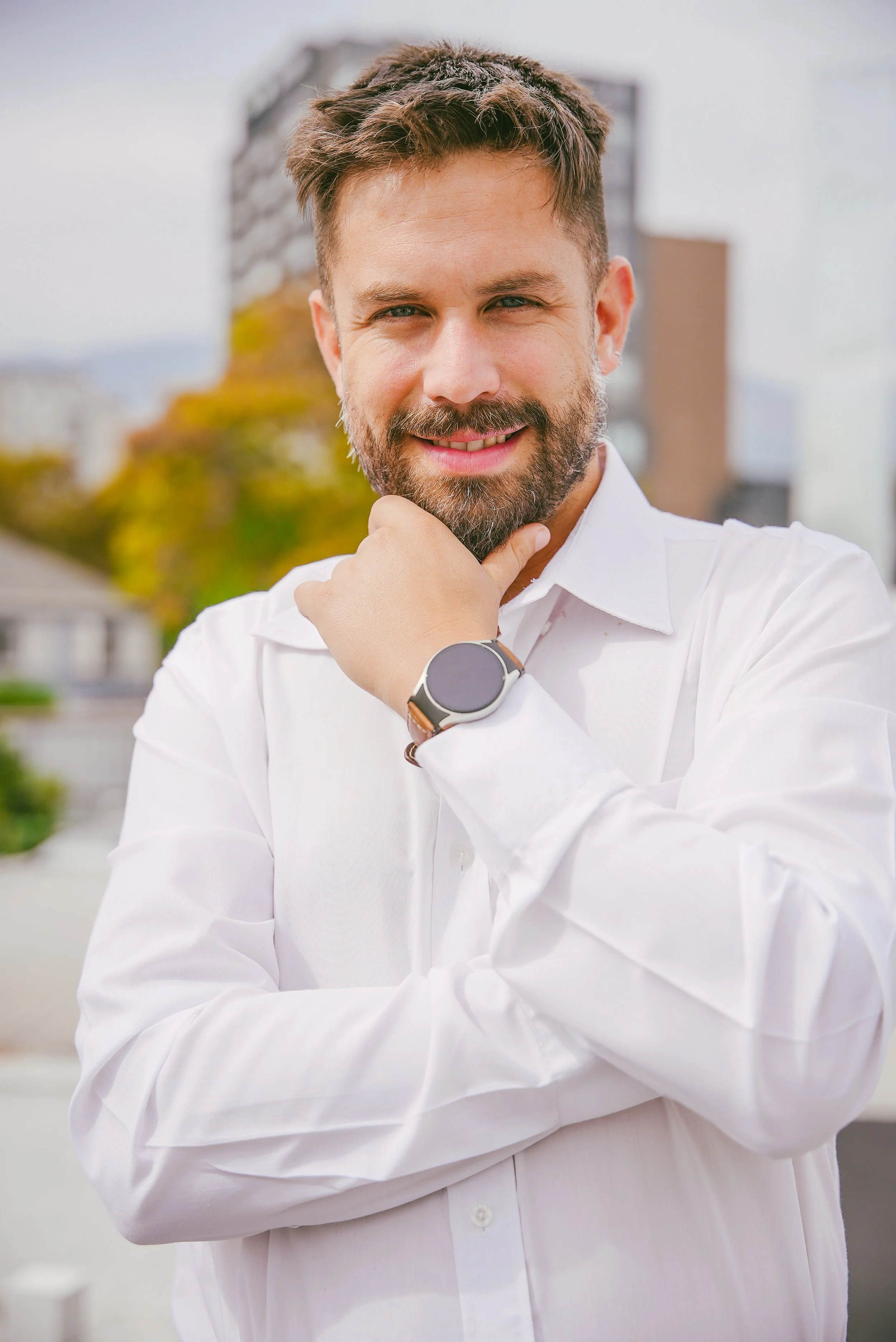 Hombre con barba, camisa blanca y reloj smartwatch, sonriendo y posando con la mano en la barbilla en un entorno urbano al aire libre.