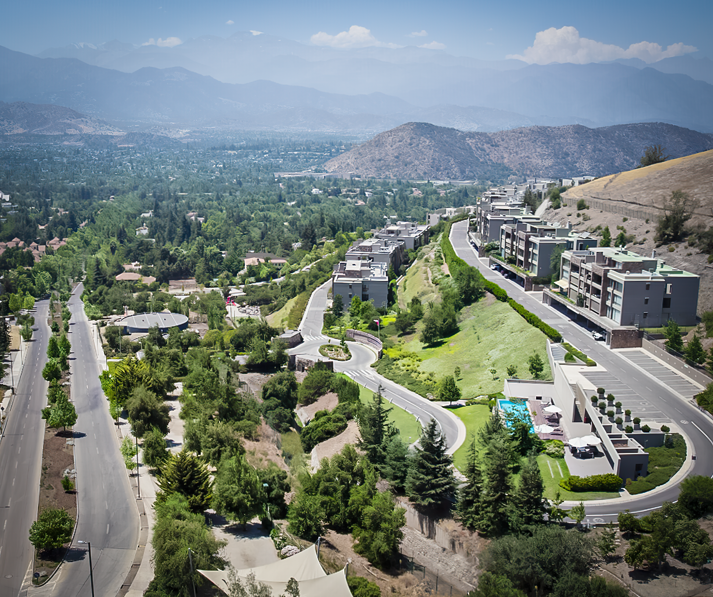 Vista panorámica de una comunidad moderna con edificios residenciales en colinas, muchas zonas verdes, árboles y una calle principal en un entorno montañoso.