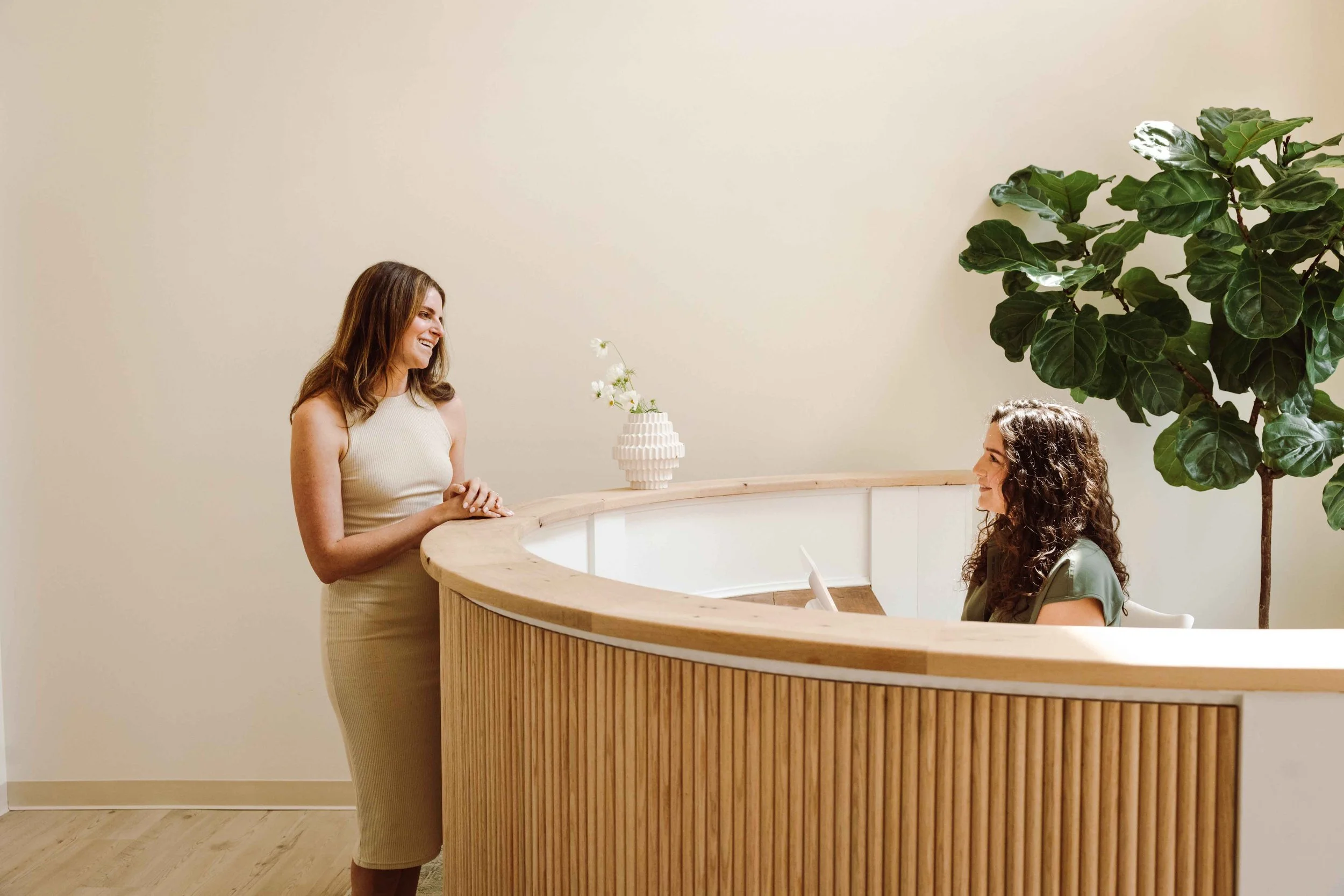 Two women are in a reception area with a curved wooden counter. One woman is standing, smiling, with her hands clasped. The other woman is seated at the counter, looking up at the standing woman. There is a large green leafy plant and a white vase with flowers on the counter.