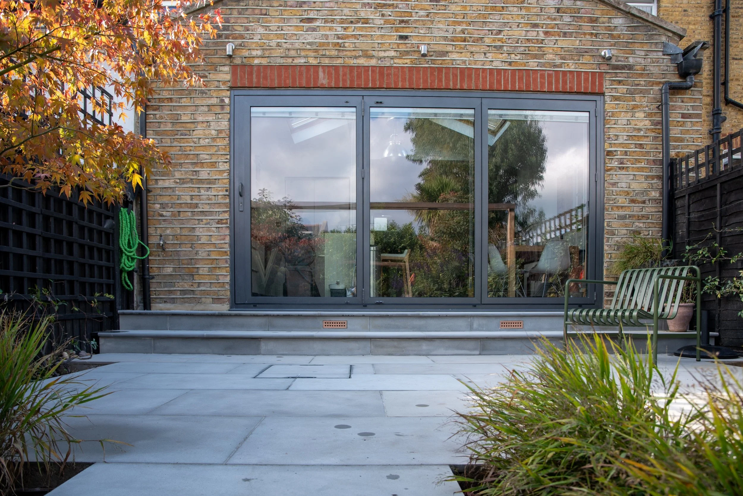 Pale grey paved patio and steps leading to bifold doors, with autumn acer and green metal bench in a Wimbledon back garden by Vlad the Landscaper