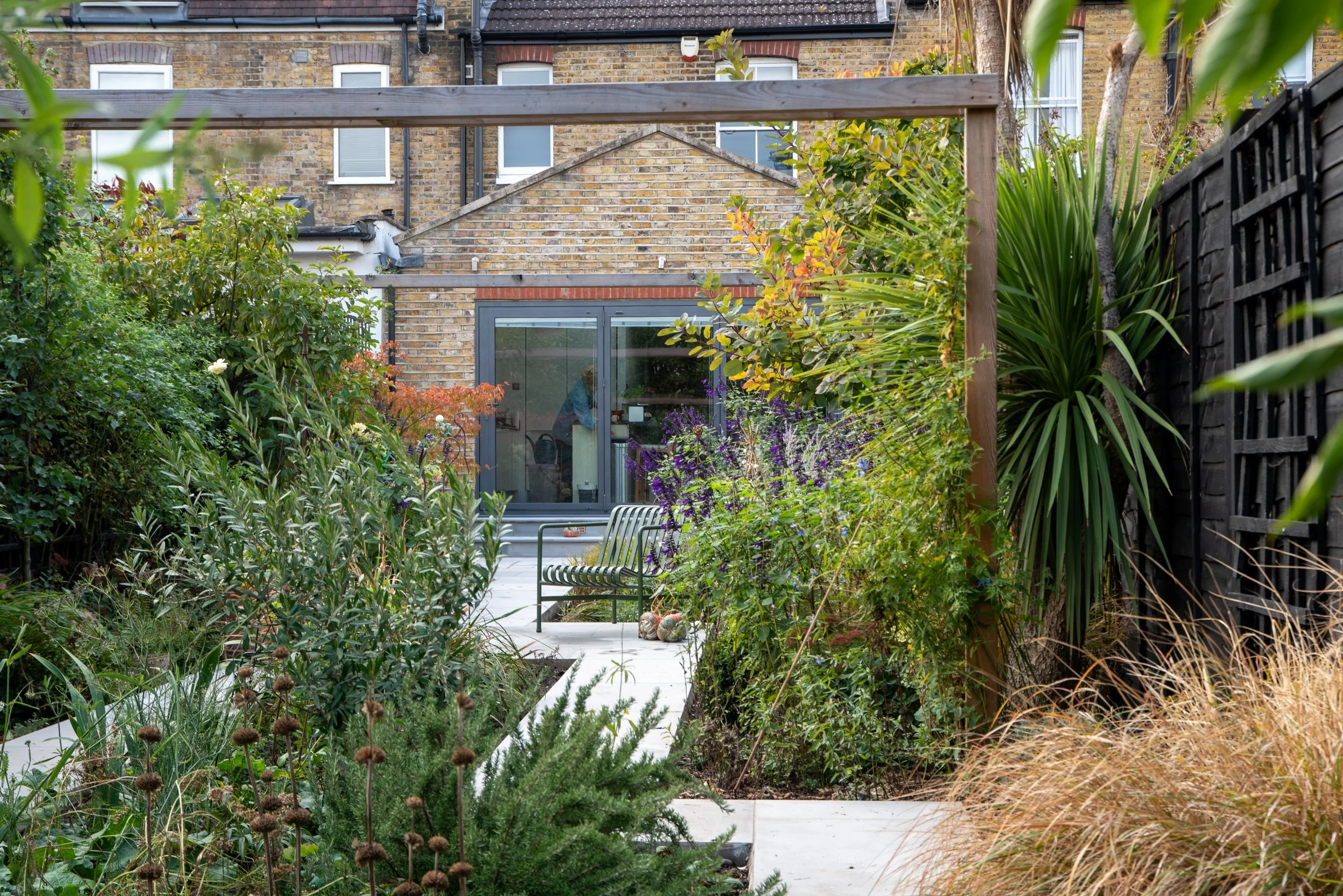 View through a timber pergola and lush planting to the house and paved terrace in a Wimbledon back garden by Vlad the Landscaper