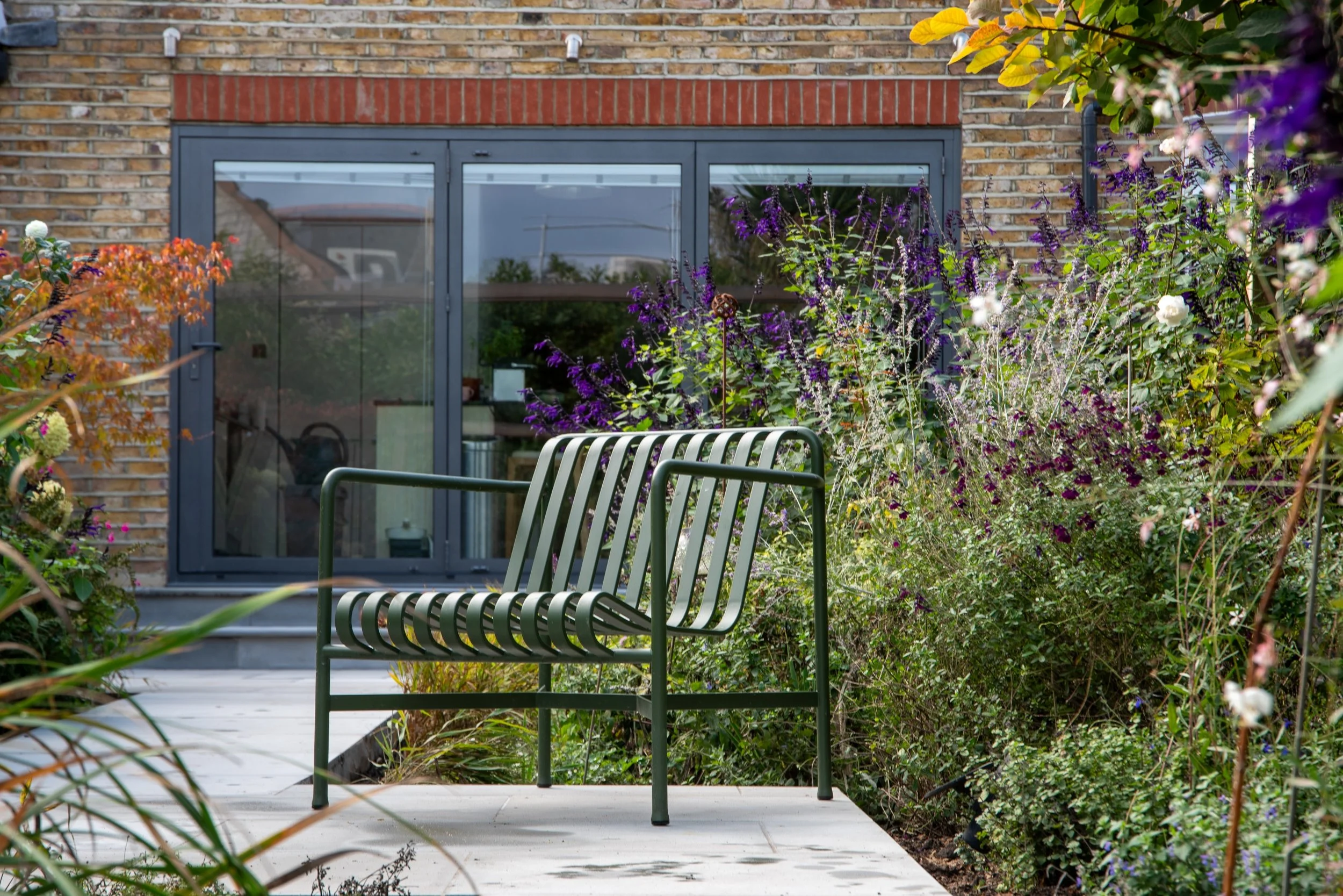 Green metal armchair on a paved terrace surrounded by purple salvia and white roses in a Wimbledon back garden by Vlad the Landscaper