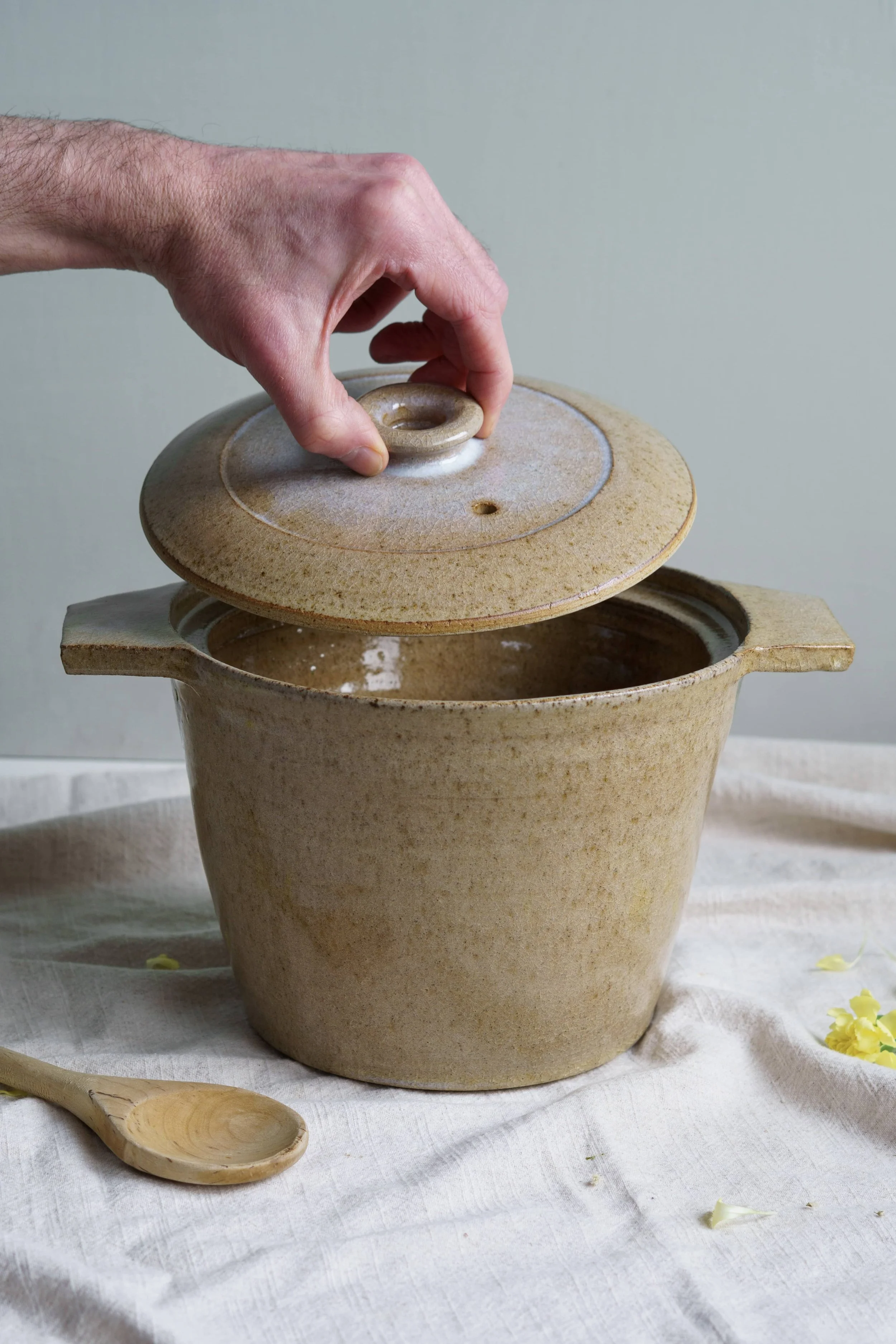 A hand lifting a ceramic lid off a casserole pot on a white cloth, with a wooden spoon nearby and yellow flower petals scattered on the cloth.