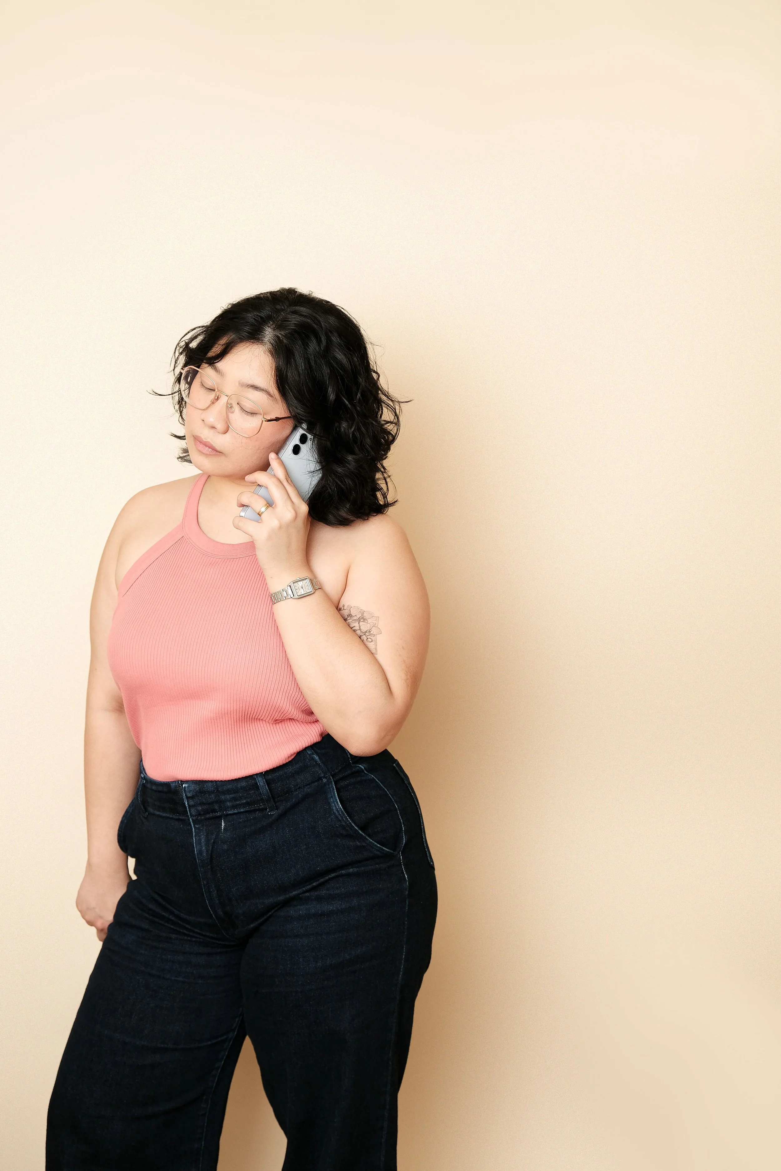 Woman with short curly black hair and glasses talking on the phone against a plain beige background.