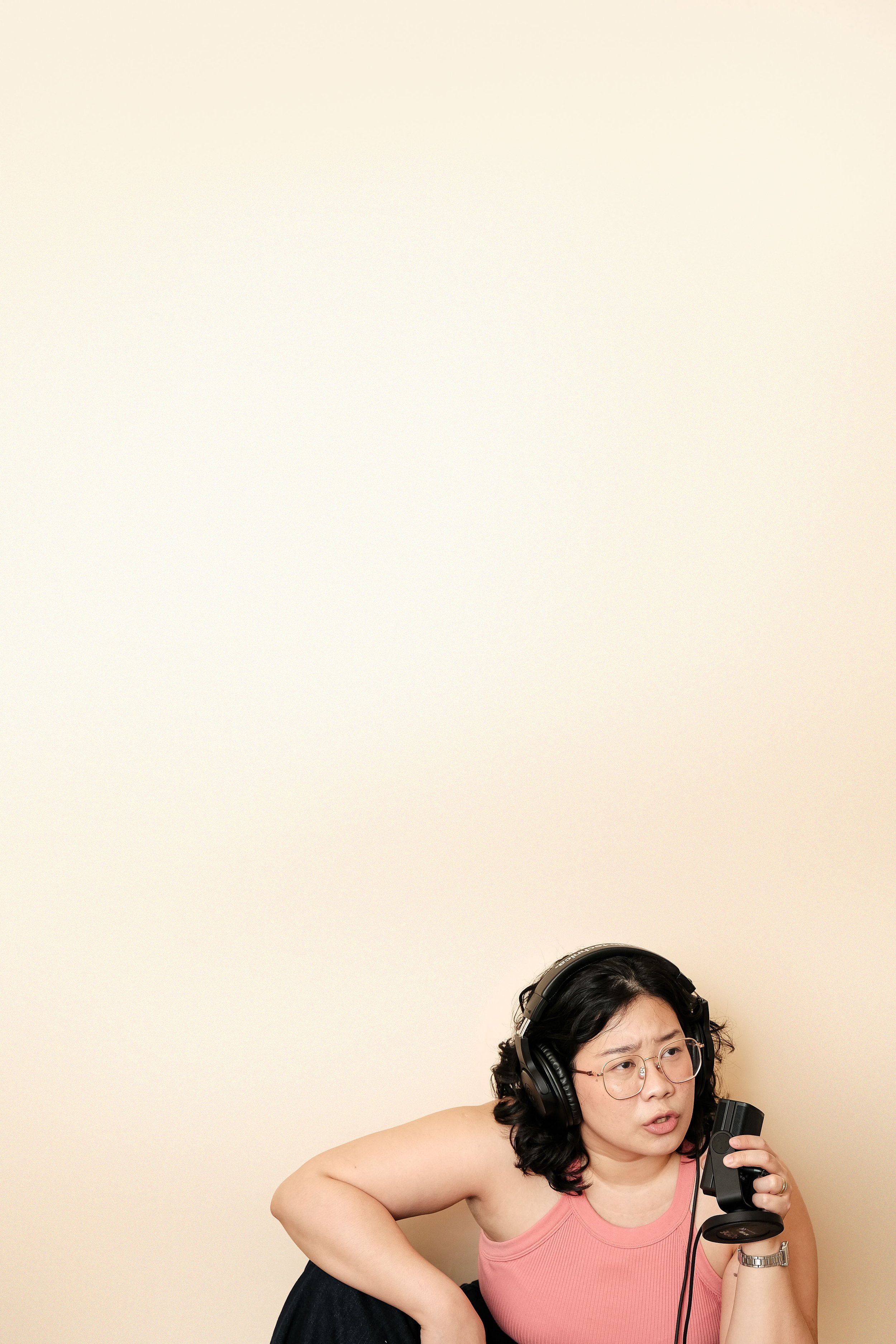 A woman with glasses and curly hair wearing headphones and a pink sleeveless top, holding a vintage microphone, looking puzzled or confused, sitting against a plain beige wall.