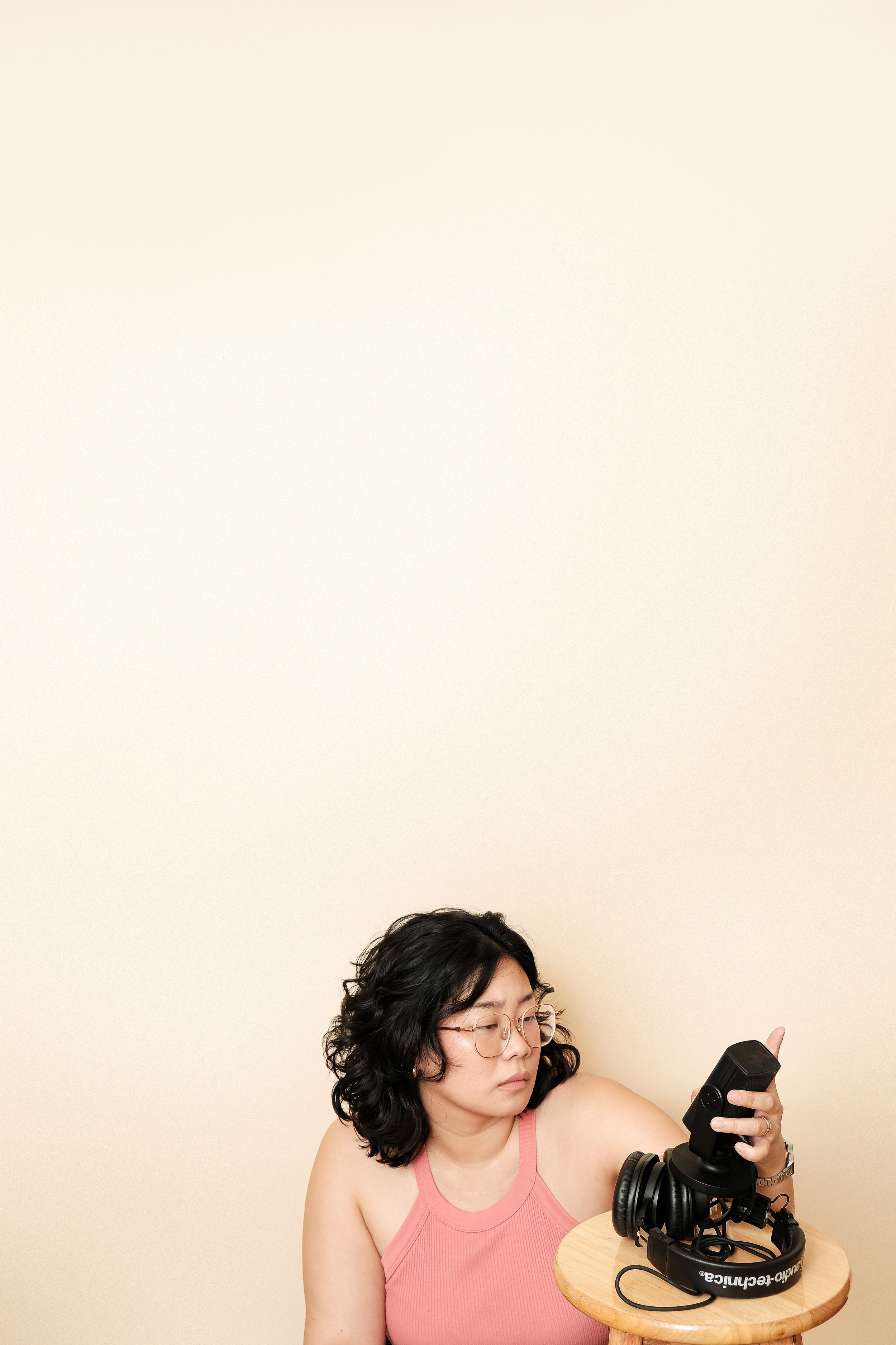 A woman with glasses and curly black hair wearing a pink sleeveless top, sitting at a small round table with headphones and a wireless microphone in her hand, against a plain beige wall.