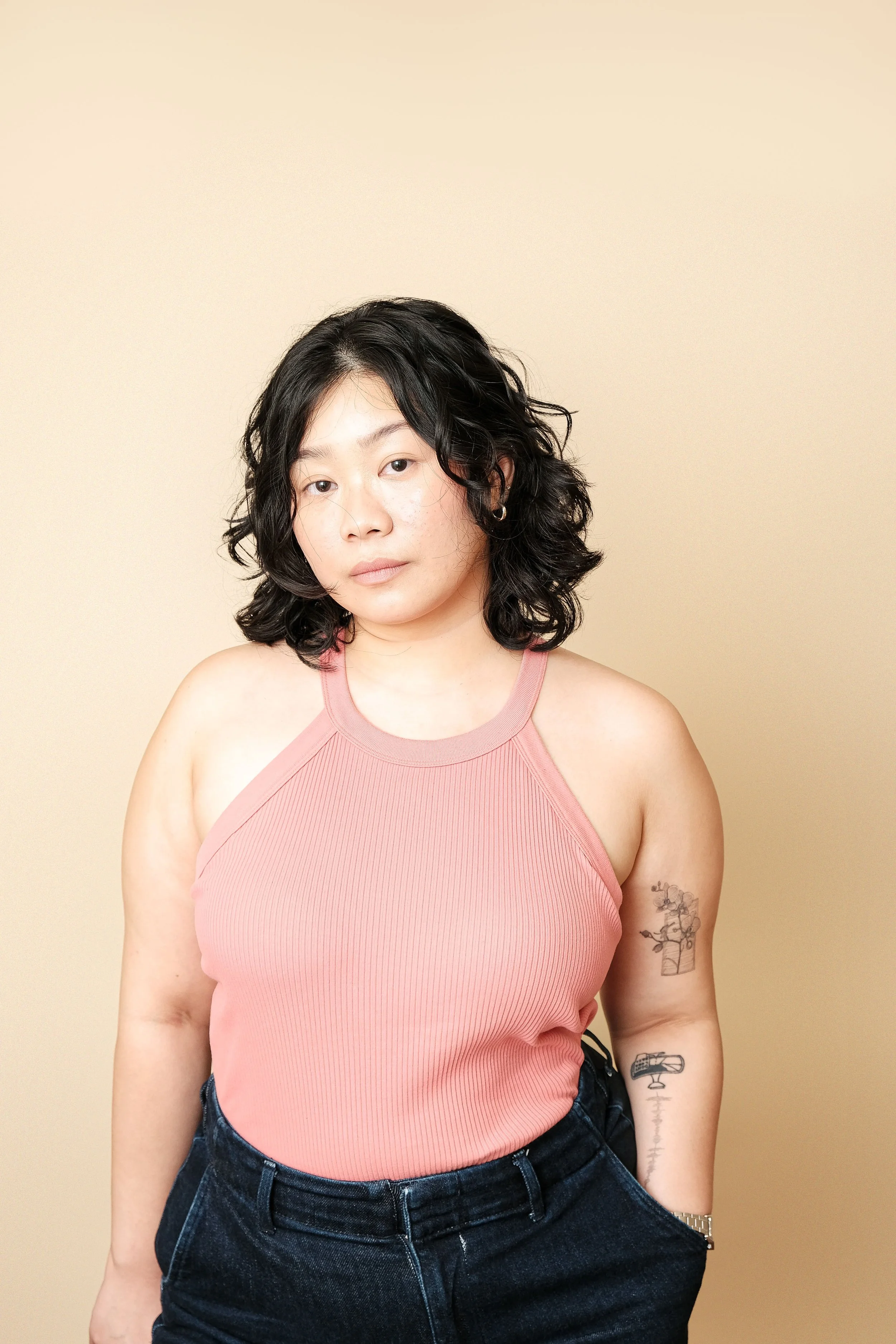 A woman with black curly hair wearing a pink sleeveless top and dark jeans standing against a beige background.