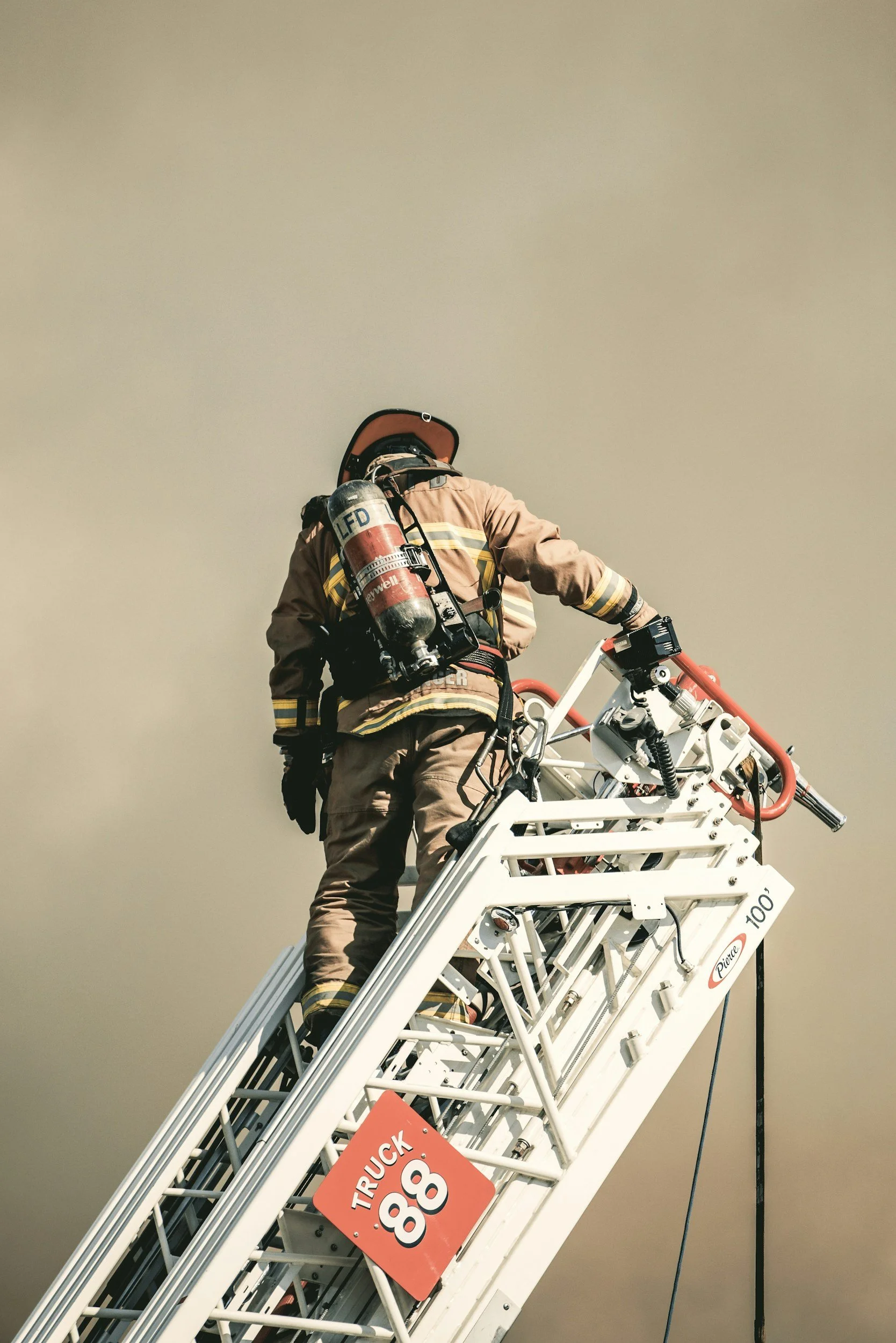 A firefighter wearing a bright yellow and black helmet and a brown uniform with reflective stripes is standing on a tall, white fire truck ladder. The firefighter has a compressed air tank backpack and is holding onto the ladder while looking down. The sky in the background appears hazy and gray.