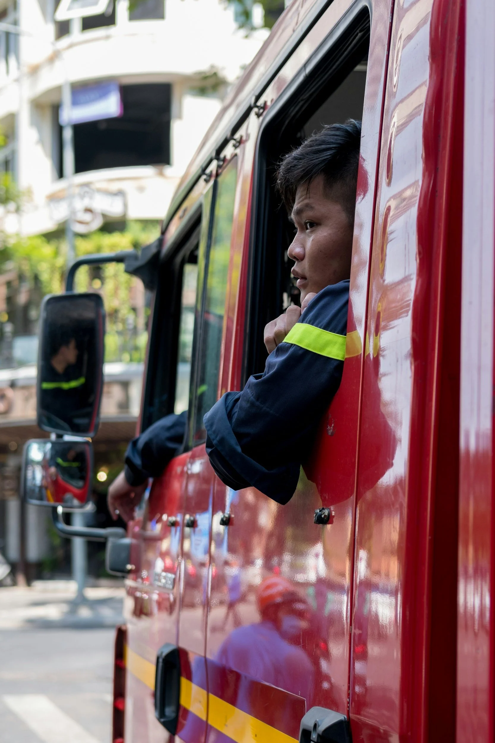 A man looks out of the window of a red emergency vehicle with a yellow stripe, parked on a city street. The background shows a blurred building and trees.