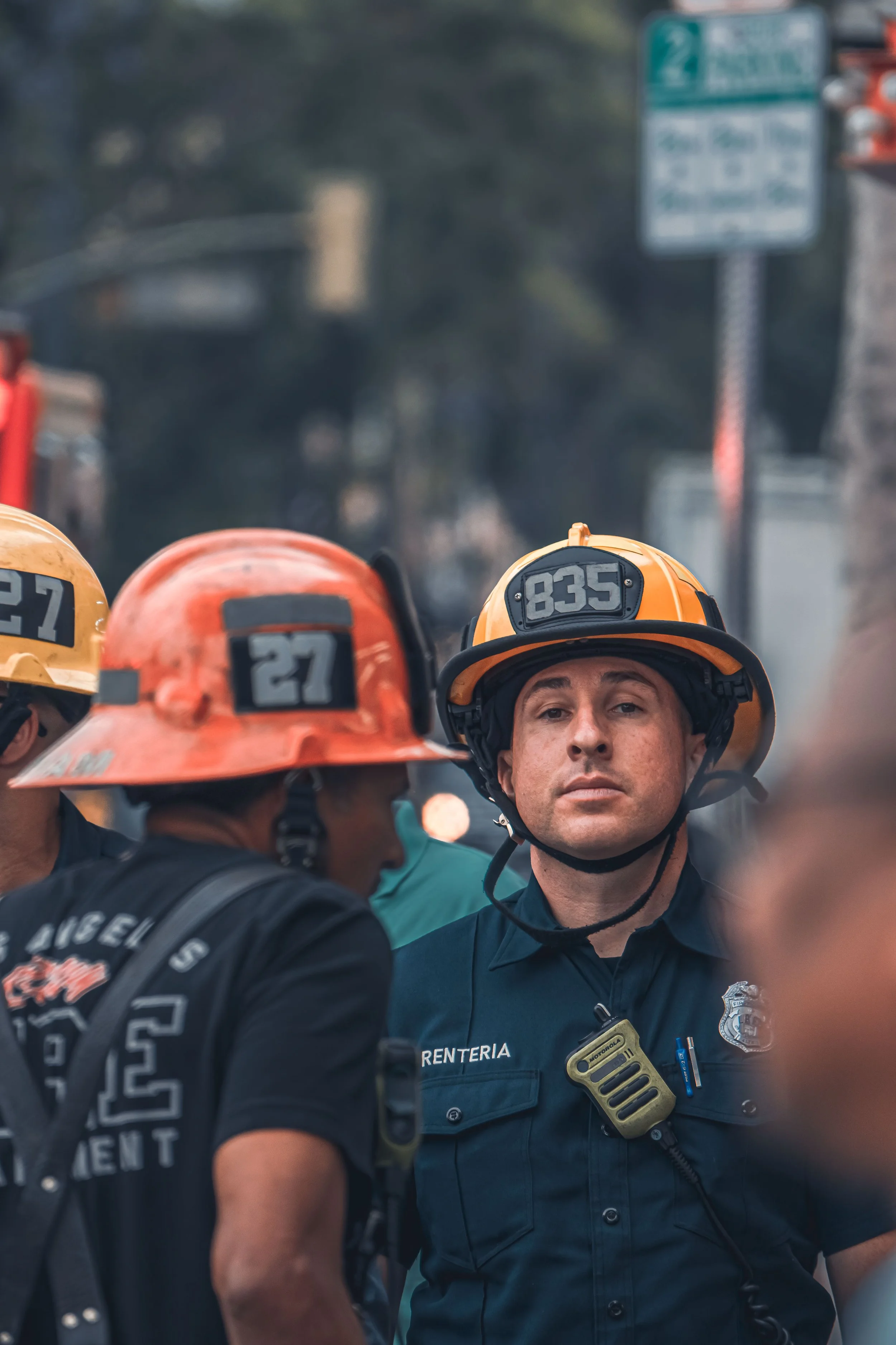 A group of firefighters, some wearing orange helmets, gathered around and listening to a firefighter in a dark uniform with a badge and radio, with a blurred background and street signs visible.
