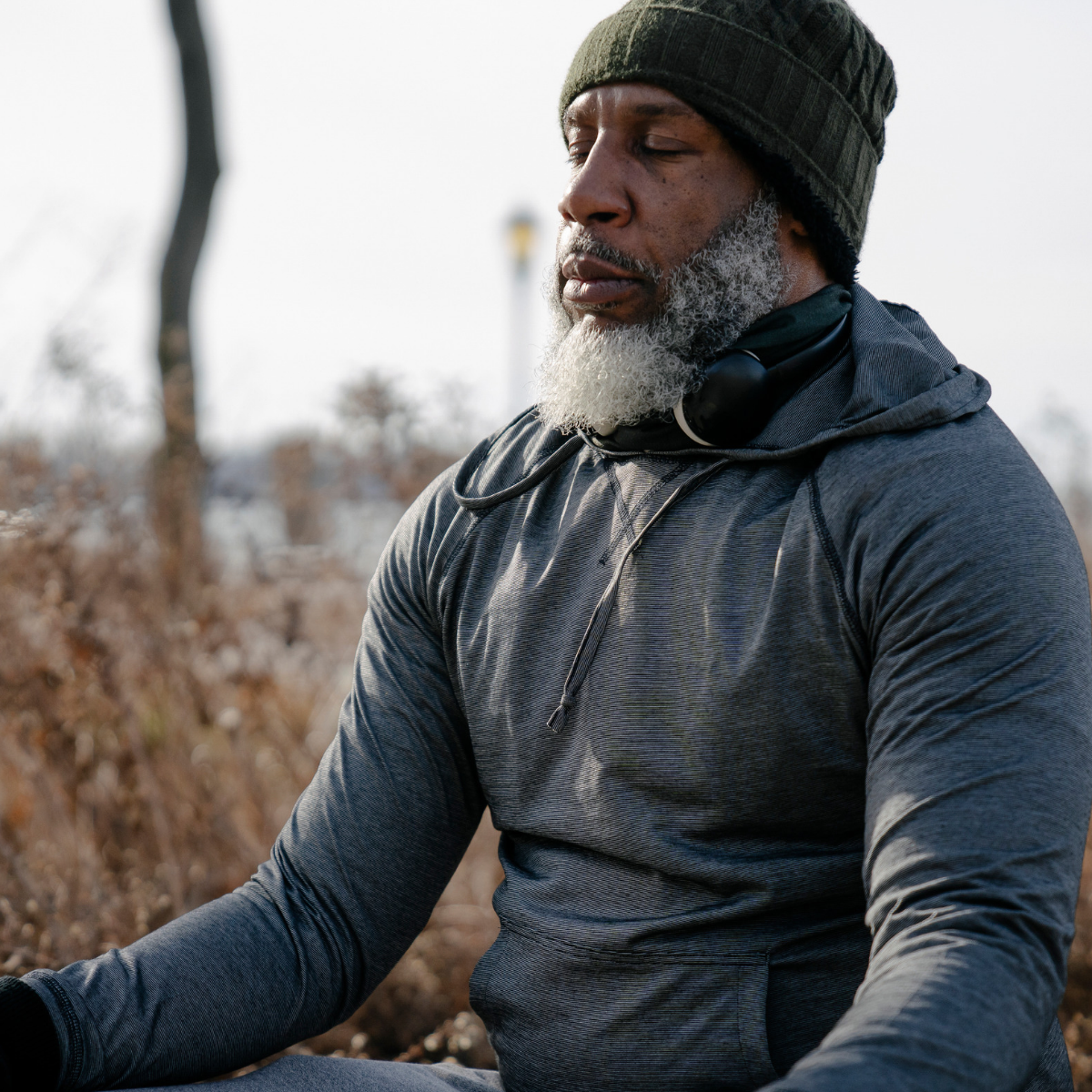 Man with gray beard meditating outdoors, wearing a green beanie, black hoodie, and headphones around neck during cloudy day.