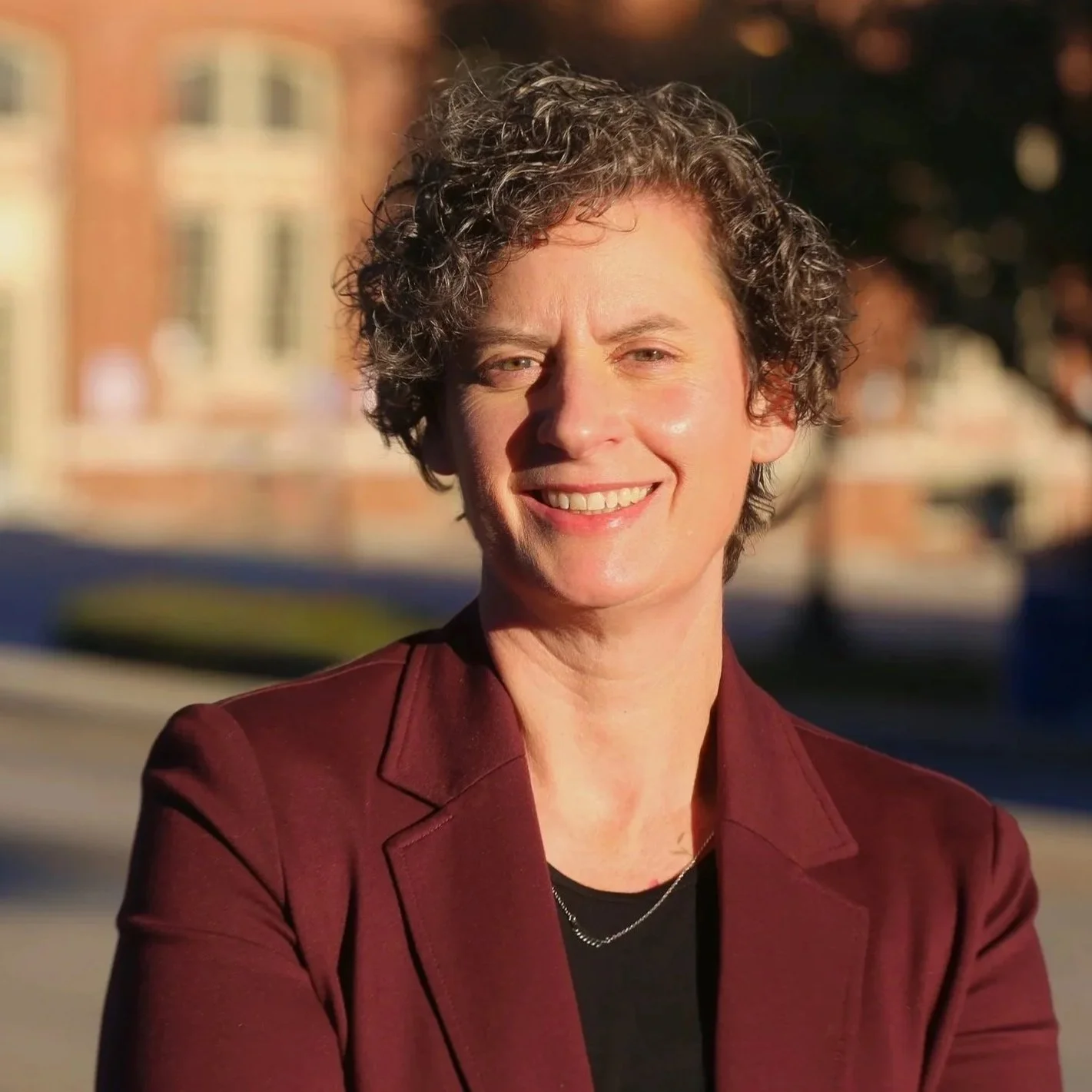 A smiling woman with short curly hair wearing a dark red blazer and a black top, standing outside in sunlight with a blurred background of buildings.