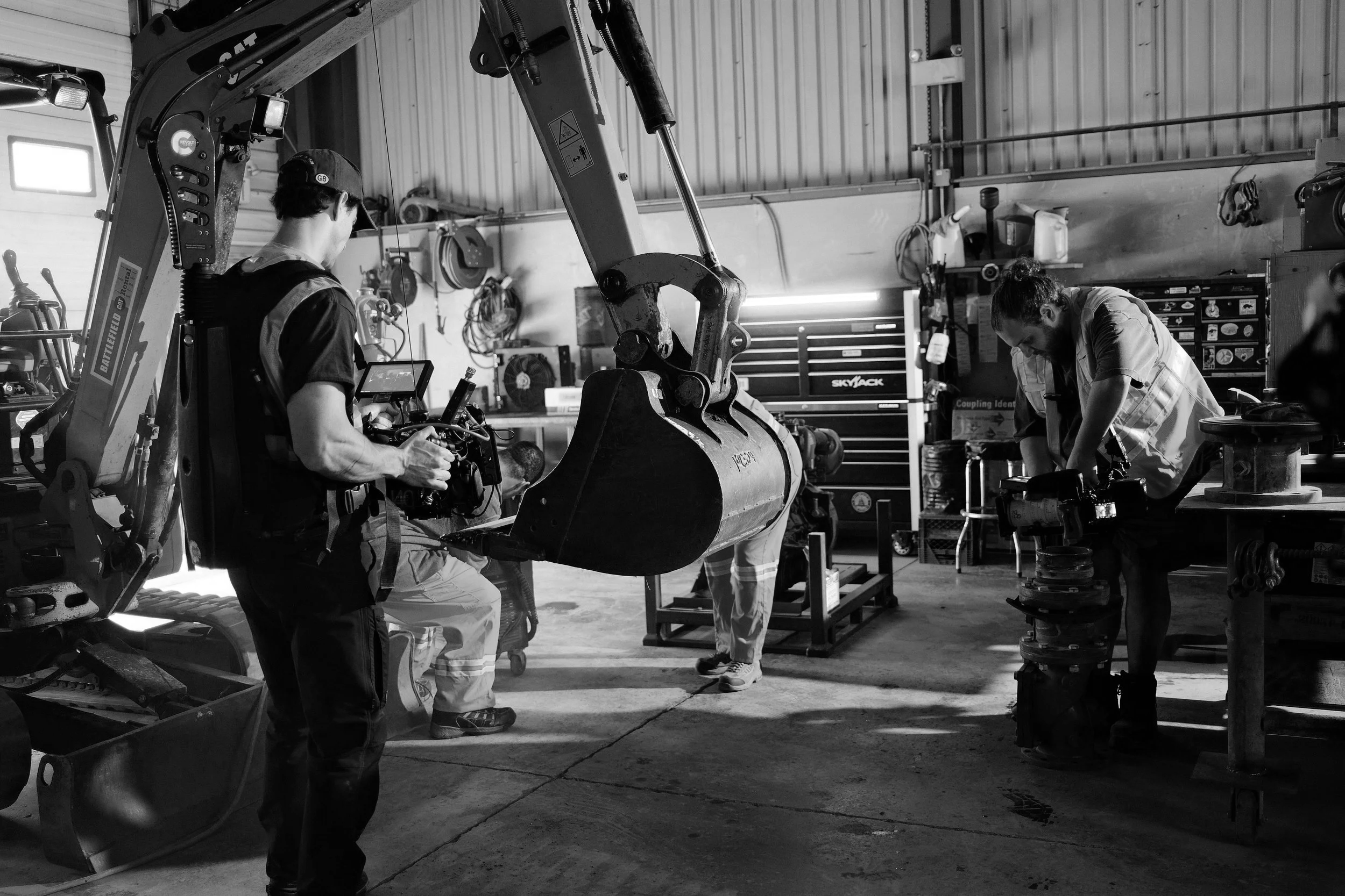 Three workers in a workshop working on a large excavator bucket. One person is filming or taking pictures with a camera.