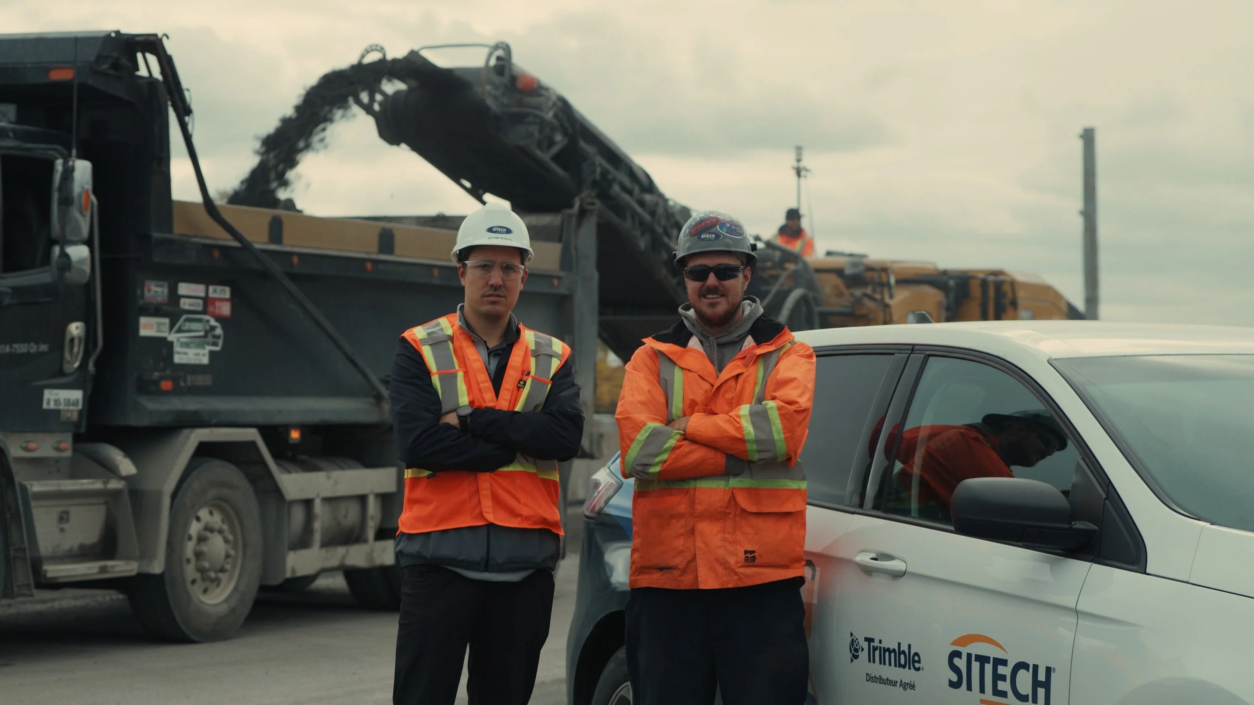 Two construction workers wearing safety vests and hard hats standing next to a white site vehicle with company logos. In the background, a road paving machine and an asphalt paver pouring asphalt, with cloudy sky overhead.