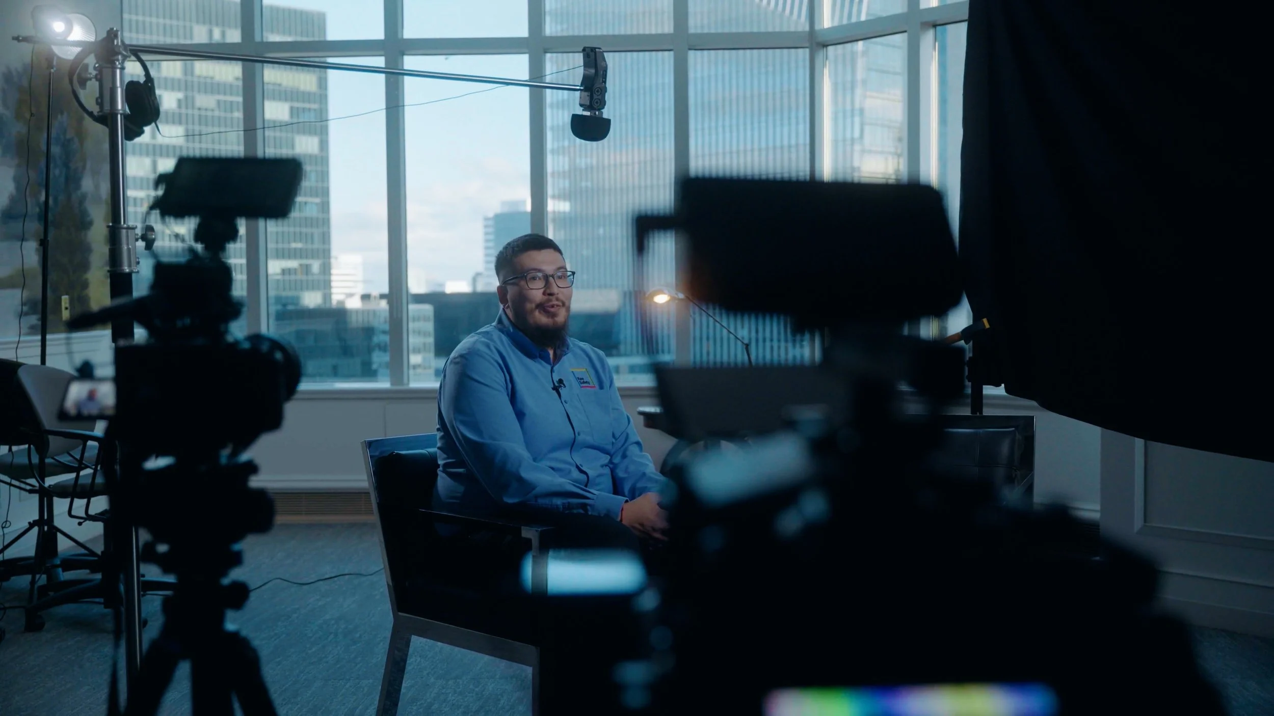 A man with glasses and a beard sitting in a chair being filmed in a studio with large windows showing city buildings, cameras, and lighting equipment around him.