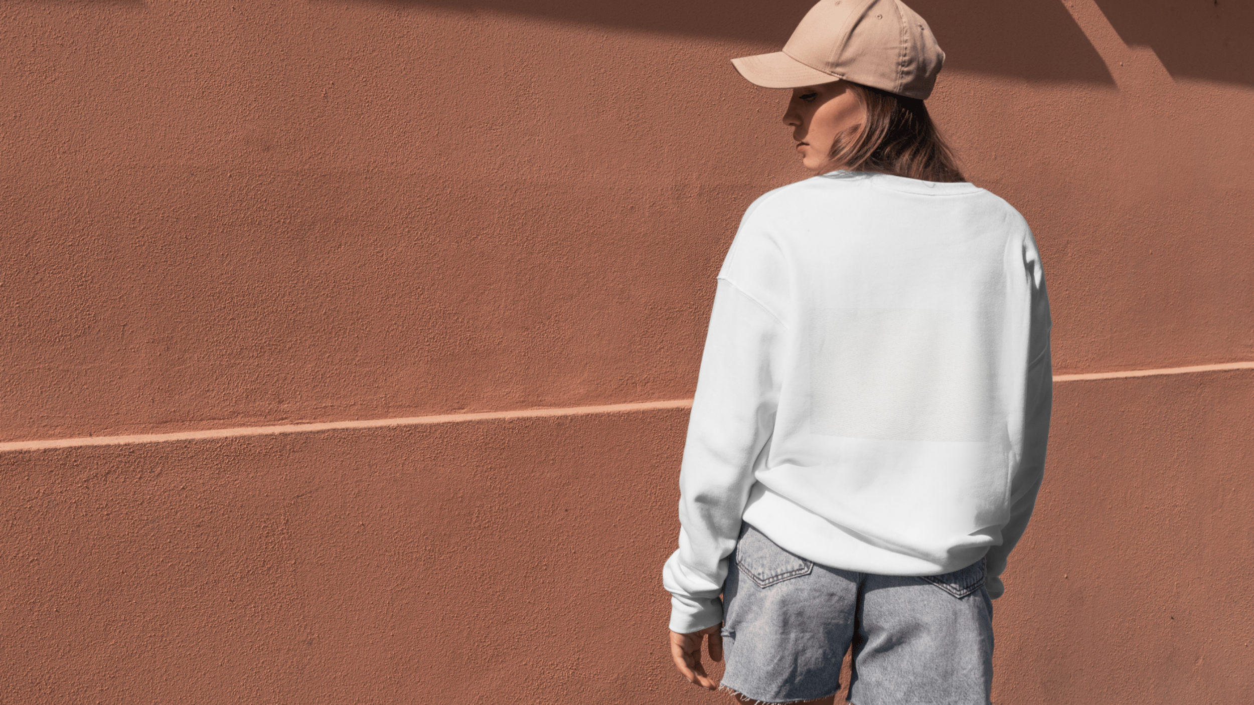 A woman with long brown hair, wearing a beige cap, a loose white sweatshirt, and gray denim shorts, standing outdoors against a textured coral-colored wall.
