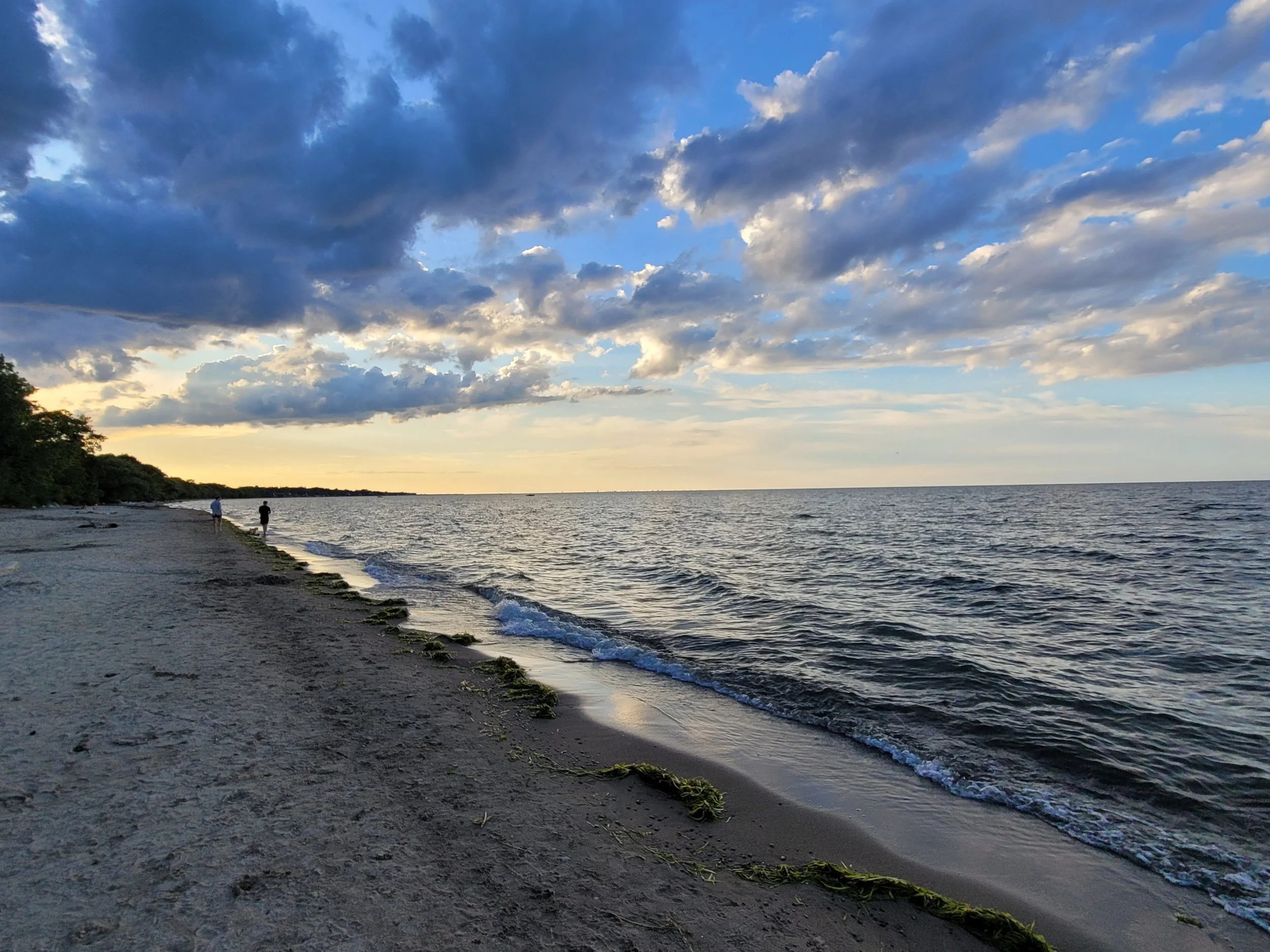 A beach at sunset with a partly cloudy sky, two people walking along the shoreline, and seaweed on the sand.