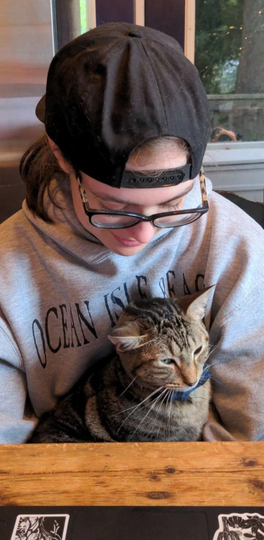 Mollie wearing glasses and a backward cap sitting with a tabby cat, Paweseidon, on their lap at a wooden table.