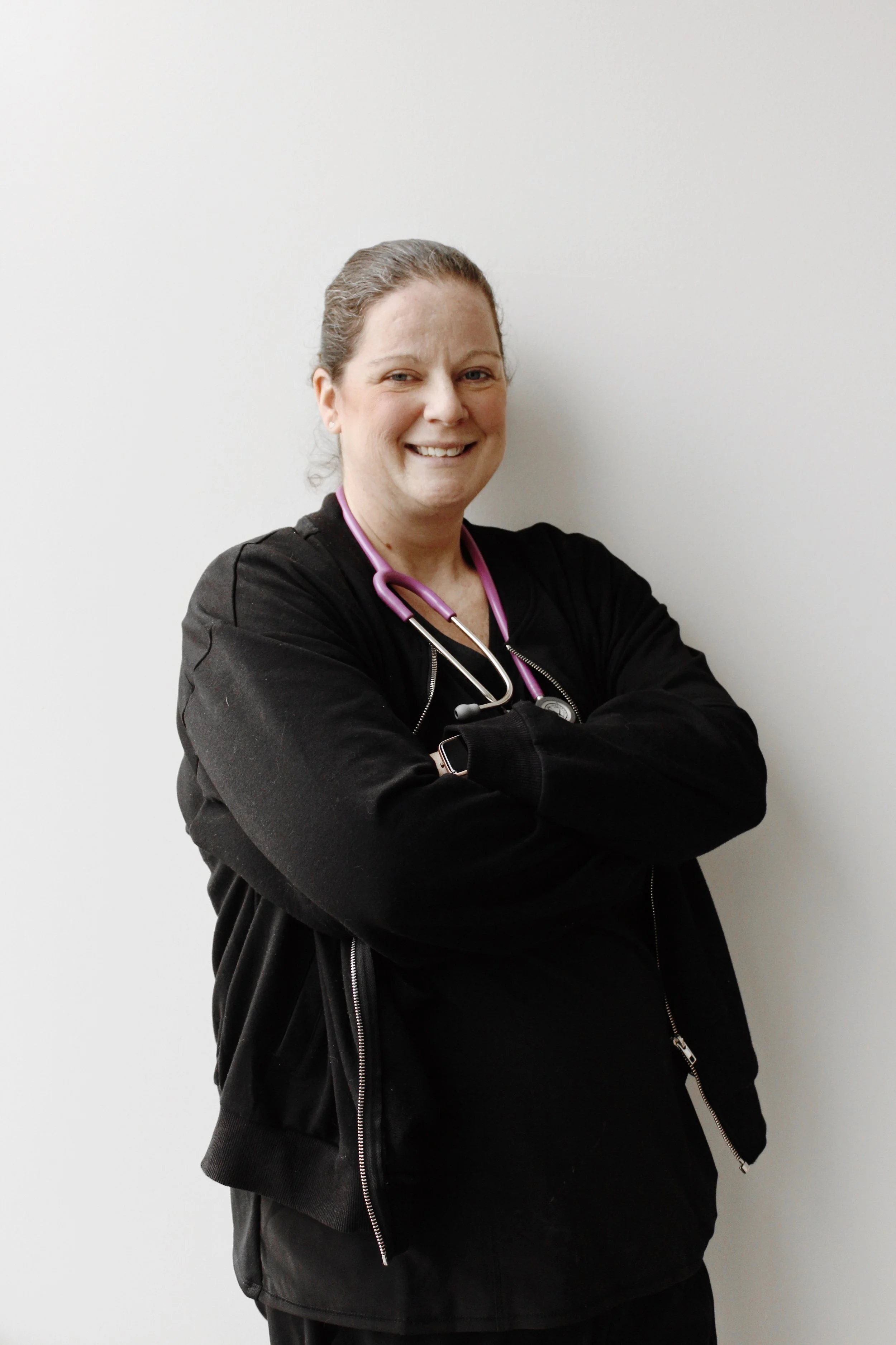 A smiling female healthcare professional with a pink stethoscope around her neck, wearing a black jacket, standing against a plain white wall.