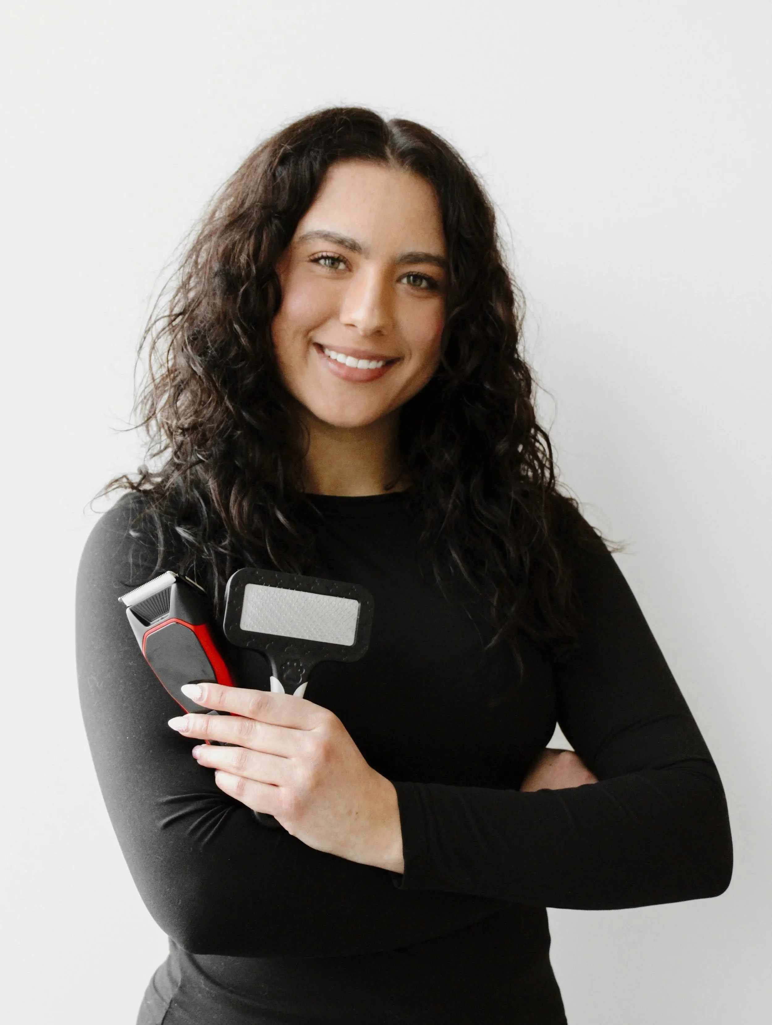 A woman with dark curly hair smiling and holding electric hair clippers and a reflection guard brush, standing against a plain white wall.