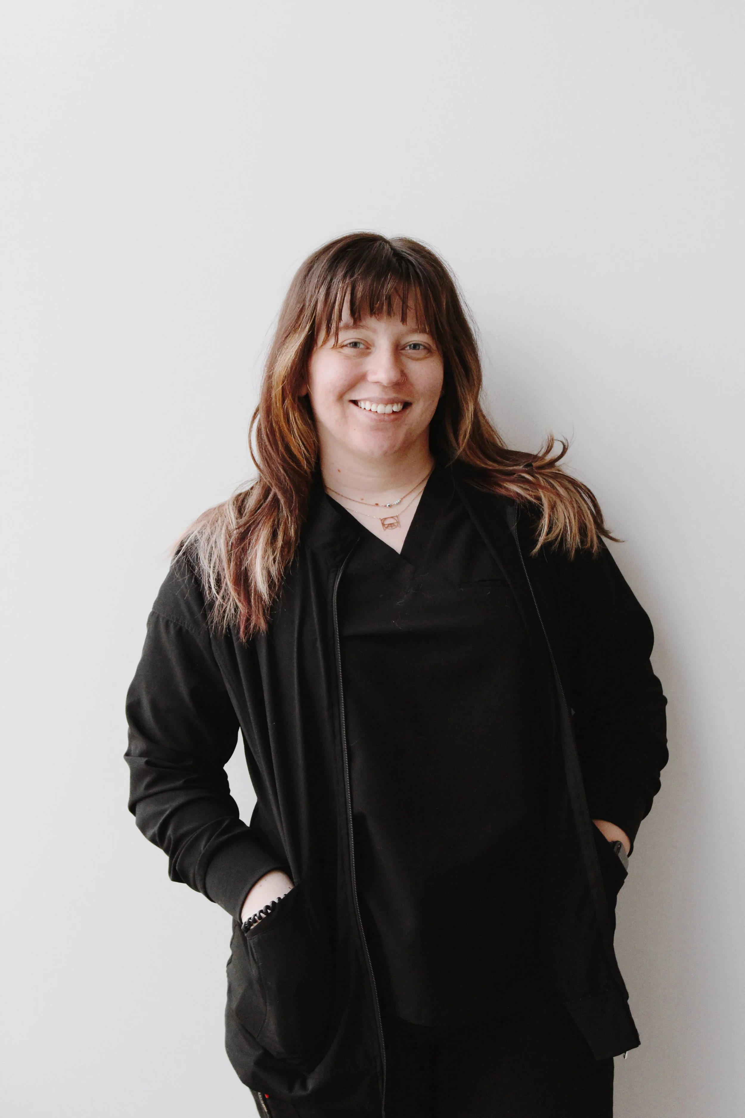 A woman with brown hair, wearing a black jacket and black top, smiling and standing against a white wall.