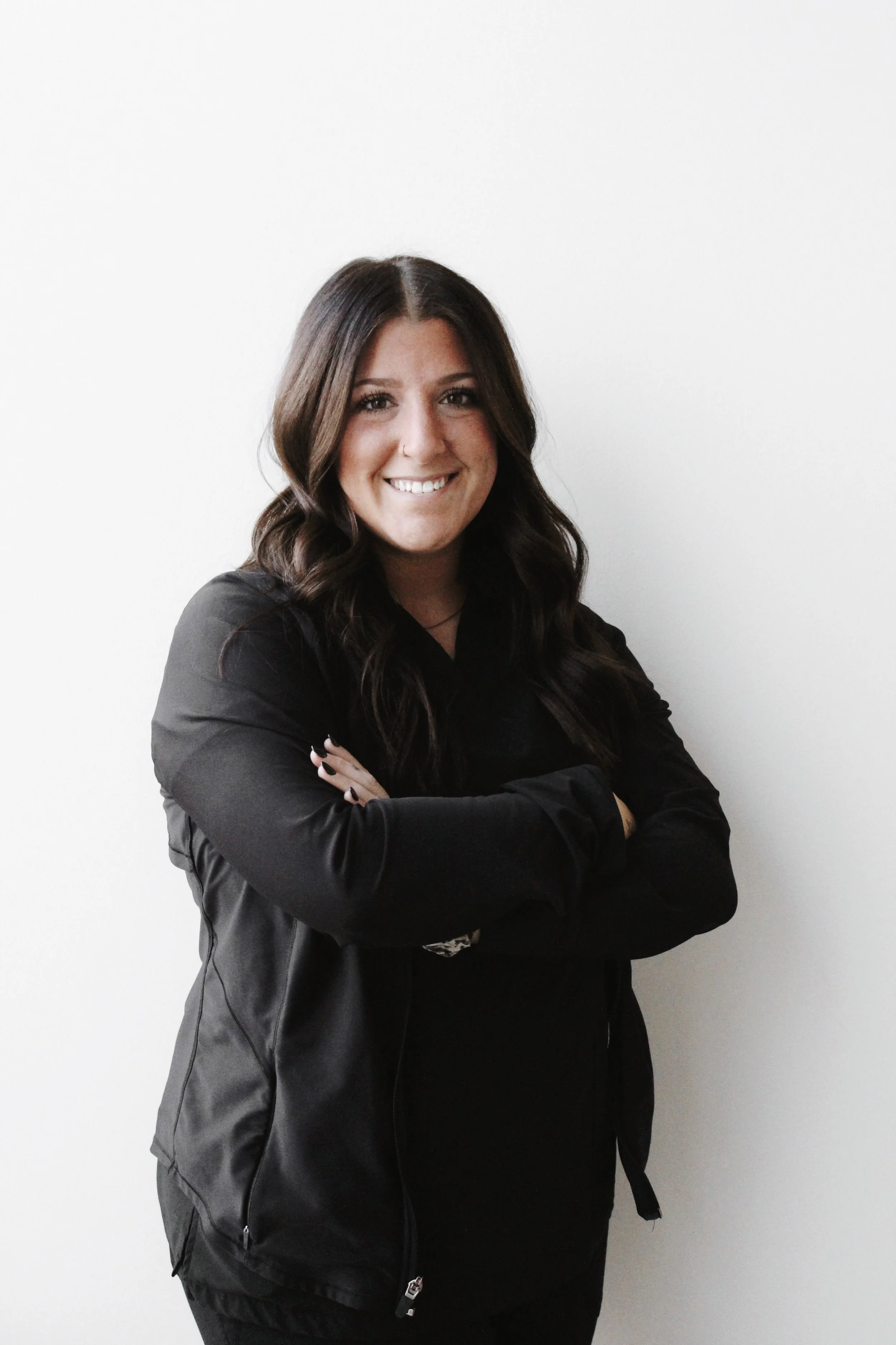 Young woman with long dark hair smiling, wearing a black jacket and top, standing with crossed arms against a white wall.