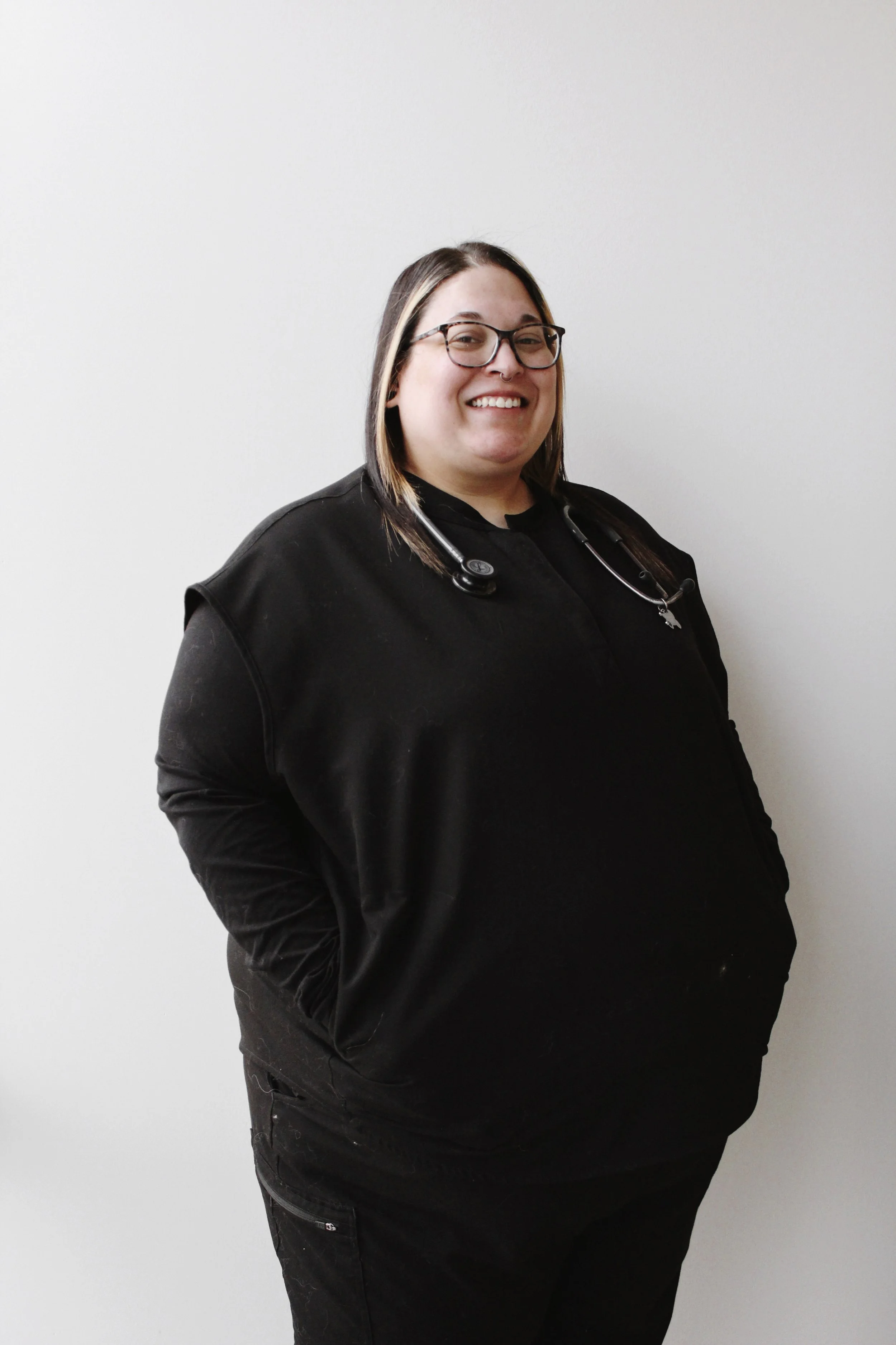 A woman with glasses, wearing a black shirt and jacket, stands against a plain white wall, smiling, with a stethoscope around her neck.