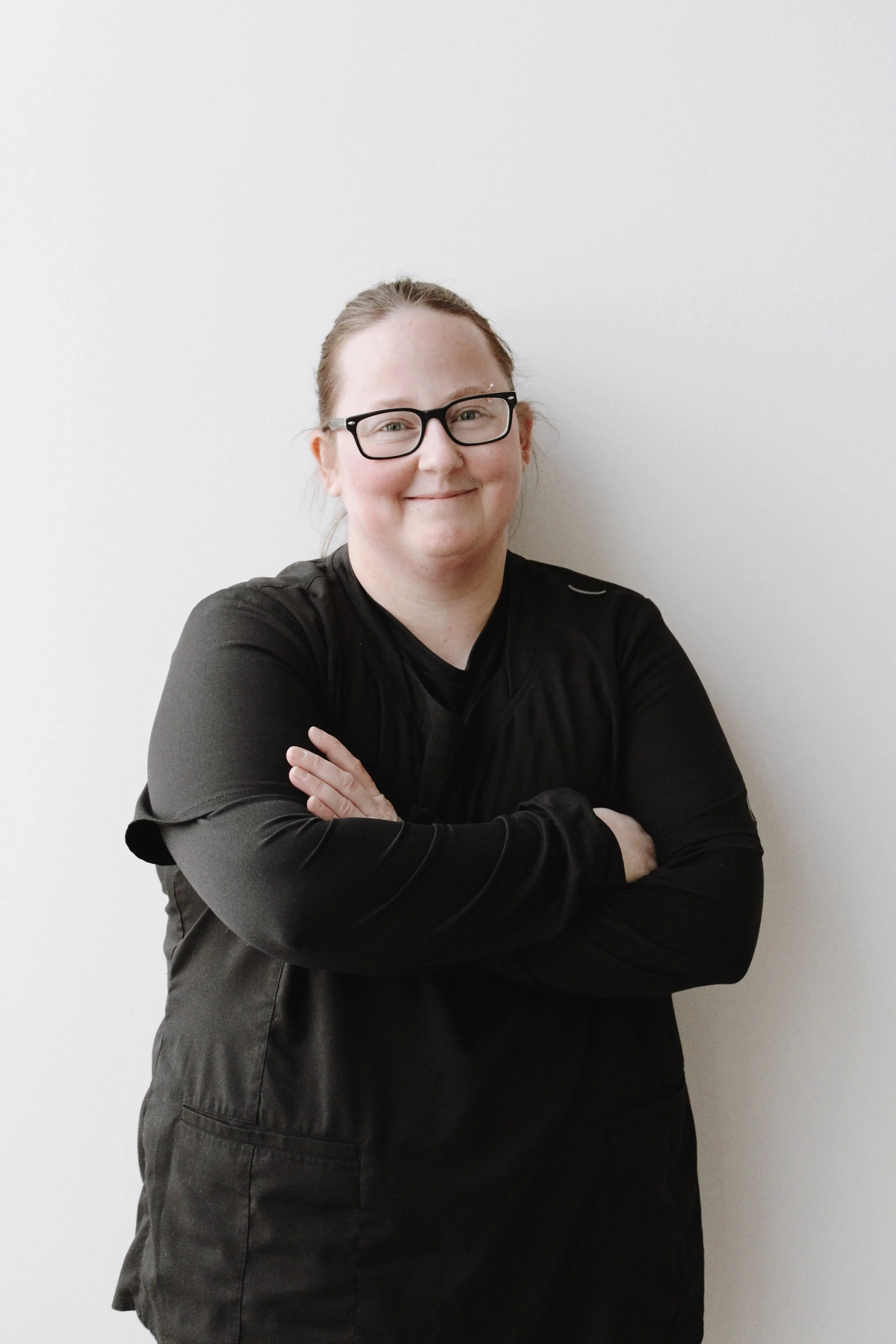 A woman with glasses and light skin, smiling with her arms crossed, wearing a black long-sleeve shirt against a plain white background.