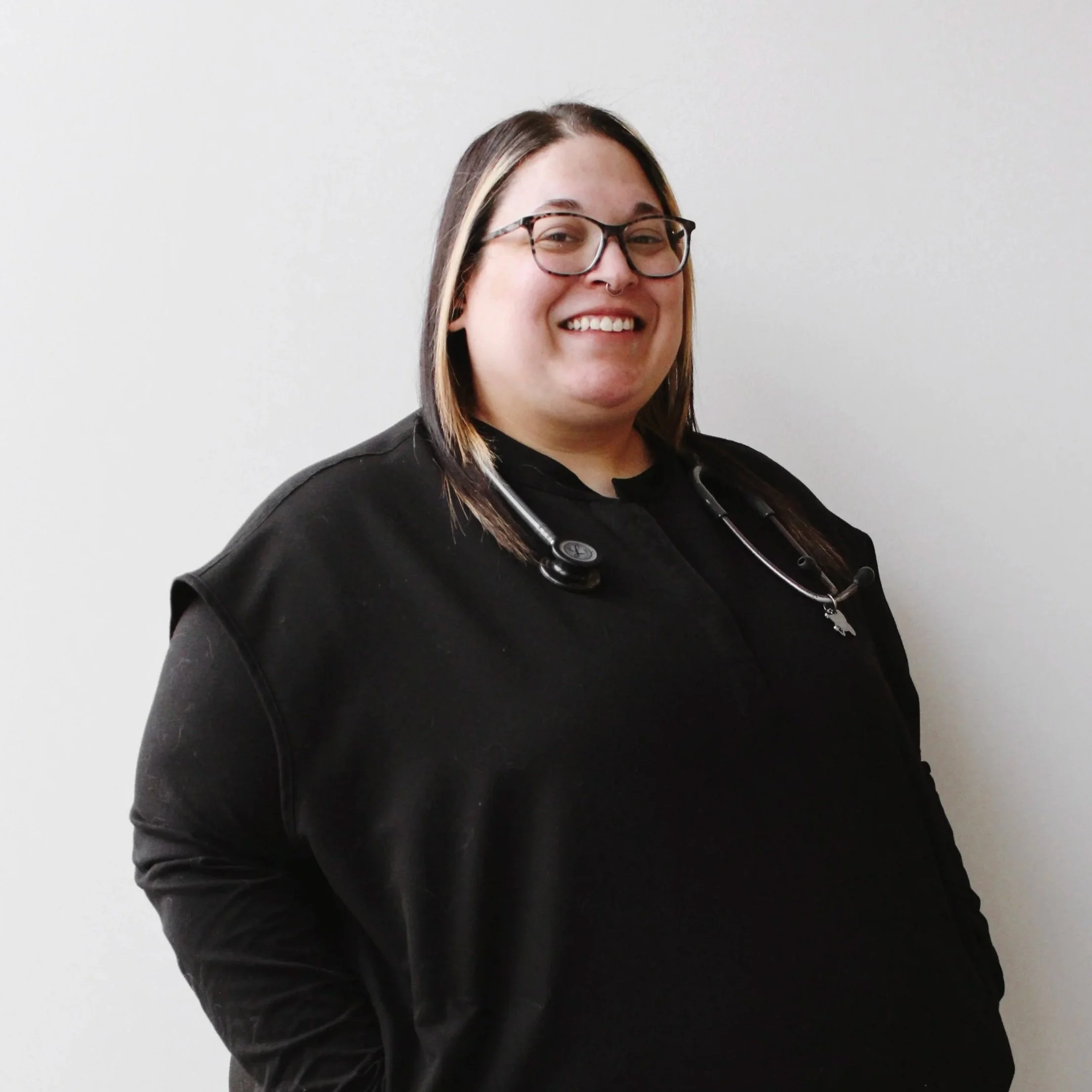A woman with glasses, wearing a black shirt and jacket, stands against a plain white wall, smiling, with a stethoscope around her neck.