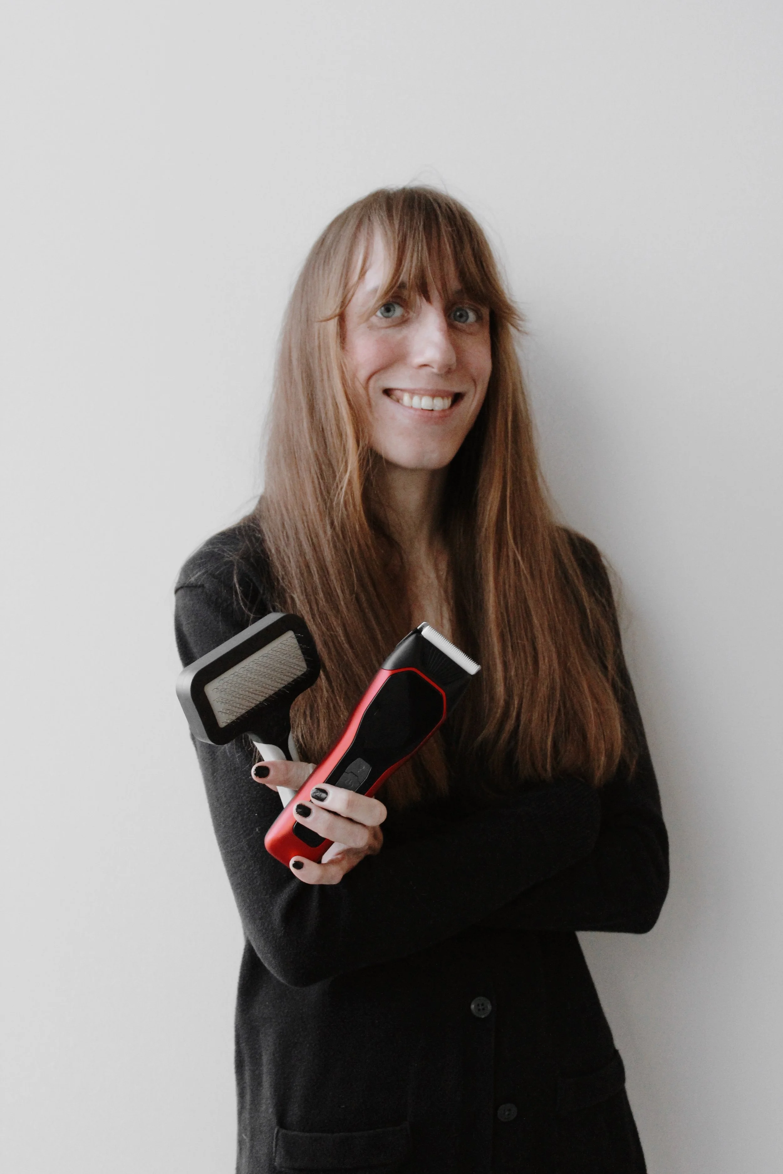 A woman with long reddish hair, smiling and holding an electric hair clipper with a brush attachment, standing against a plain white wall.
